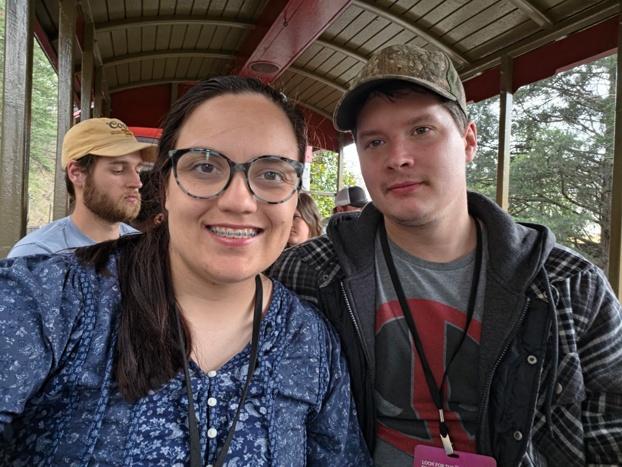 Smiling teenage girl with glasses and braces, standing next to a teenage boy with a camouflage cap, on a train or tram with other people and greenery in the background.