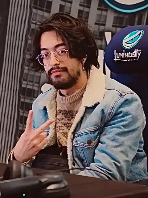 tafaar with shoulder-length hair glasses, and a beard, throwing up a peace sign while sitting in a gaming chair with the Luminosity logo. He is wearing a denim jacket over a sweater at the event Luminosity Makes Big Moves in New York, 2024.