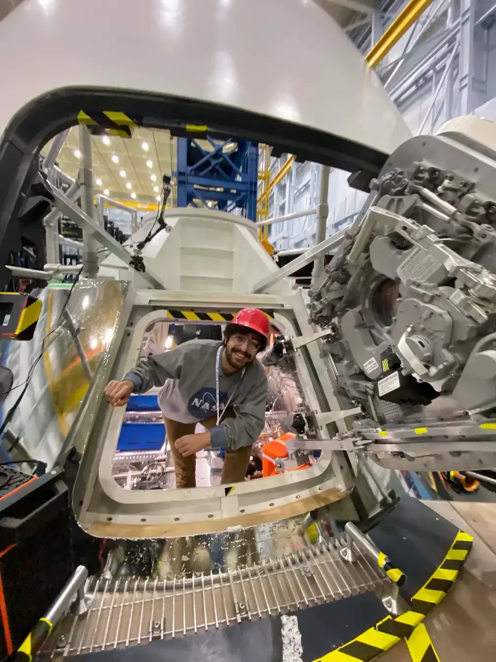 tafaar standing inside of a training module of the Orion spacecraft used on the Artemis missions at NASA. The room surrounding them is a large training facility at the Johnson Space Center.