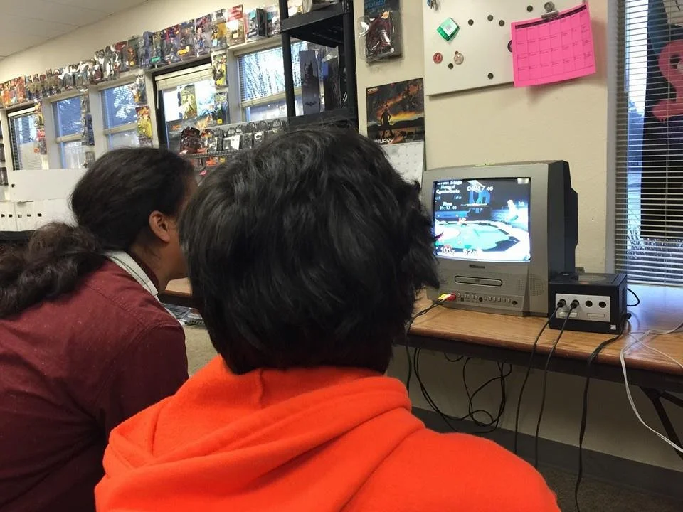 tafaar is playing Super Smash Brothers Melee on a small CRT setup. The back of his head is facing the camera. There are trading cards high on the wall in front of him, as well as a black GameCube. He is wearing an orange Goku hoodie.