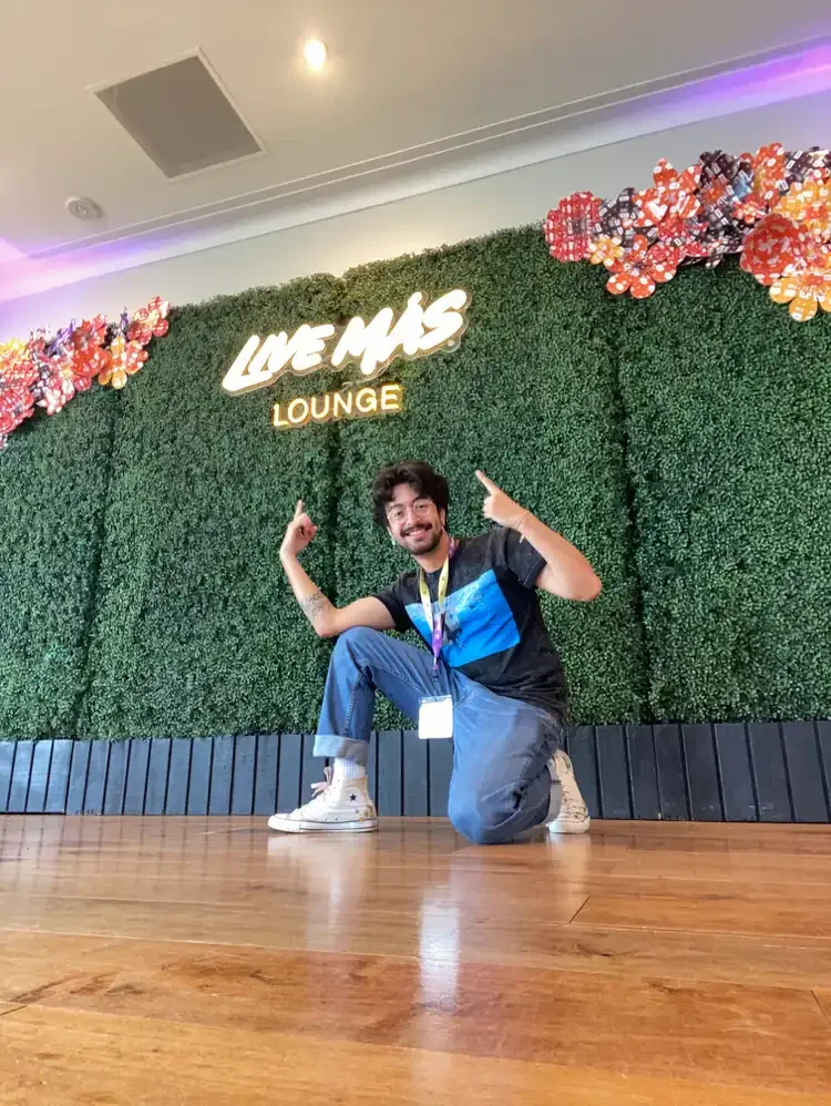 tafaar crouching on a wooden floor in front of a wall of fake hedges with colorful flowers pointing at a sign that says 'LIVE MAS LOUNGE'. This photo was taken at the Taco Bell Foundation's Summer of Connection 2025.