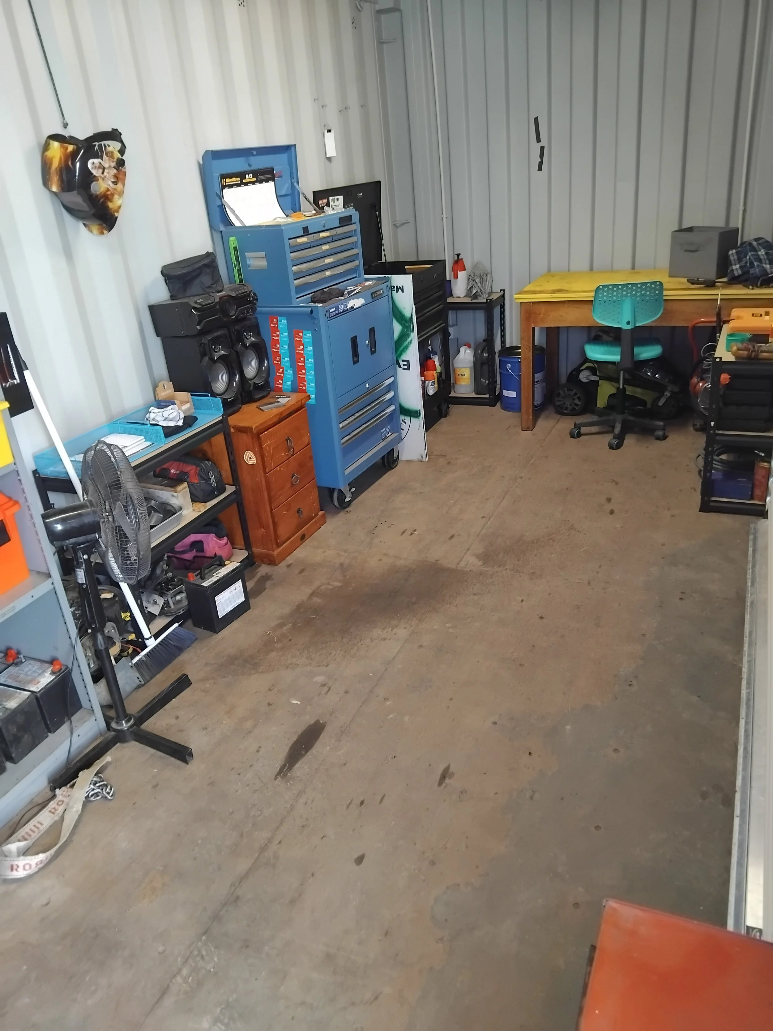 A garage or workshop space with metal walls and a concrete floor. Contains a blue tool chest, a workbench with a green chair, various tools and supplies, a speaker system, a fan, and miscellaneous items like bottles and boxes. The space appears organized but in use.