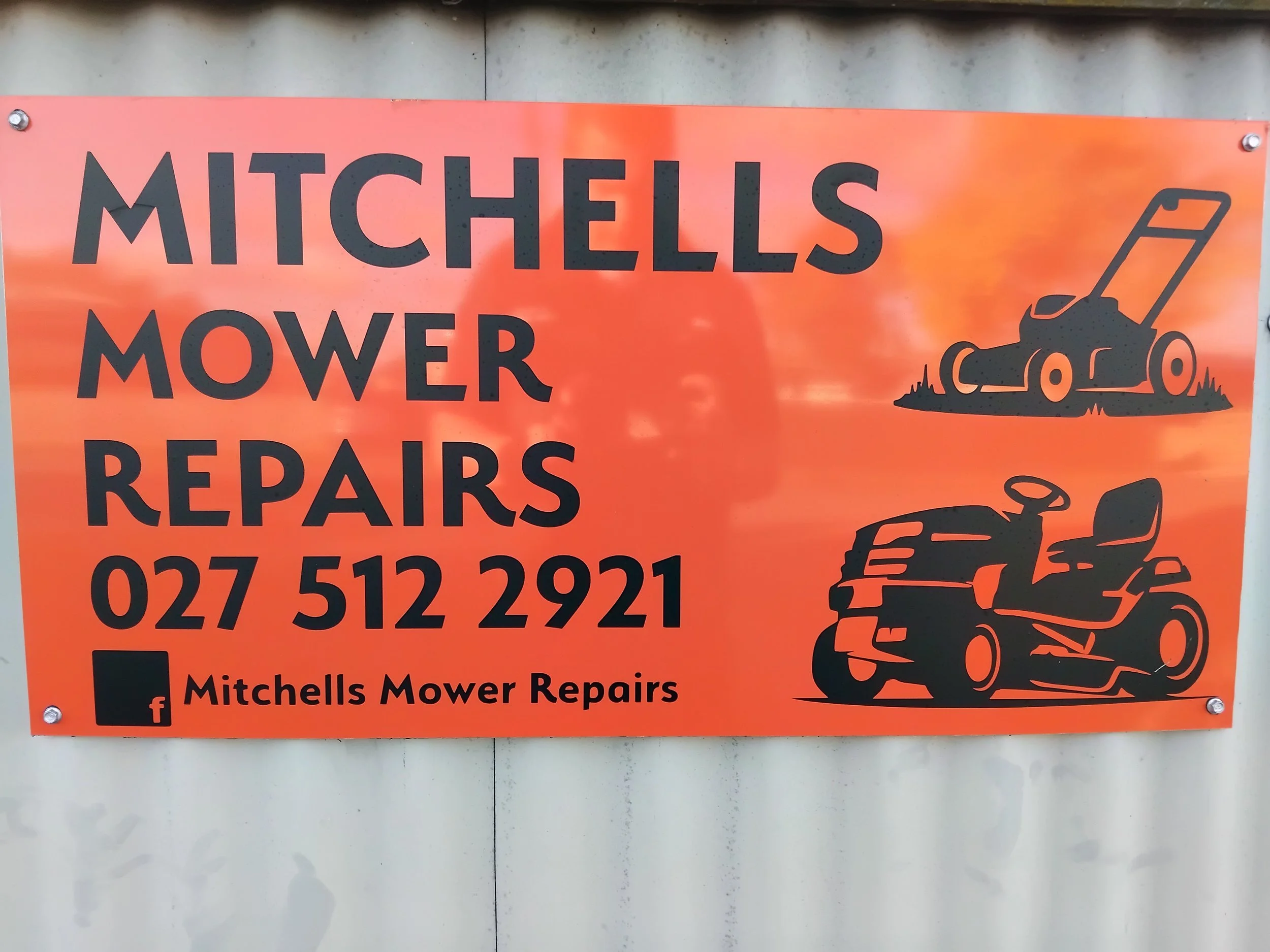 Orange sign for Mitchel's Mower Repairs with a black illustration of a riding lawn mower and a phone number, mounted on a gray wall.