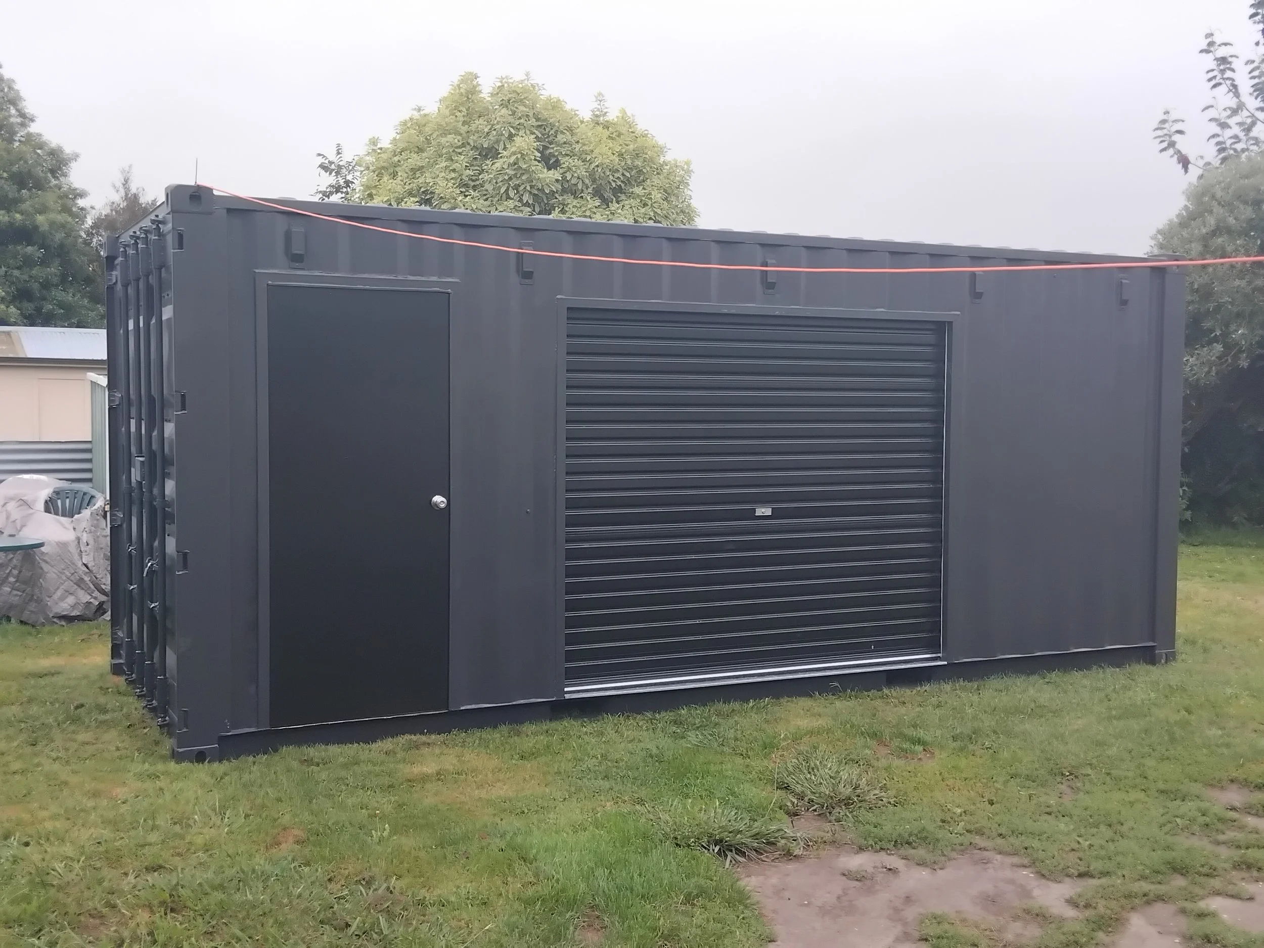 A black storage shed or container with a roller shutter door, set on a grassy area with trees and cloudy sky in the background.