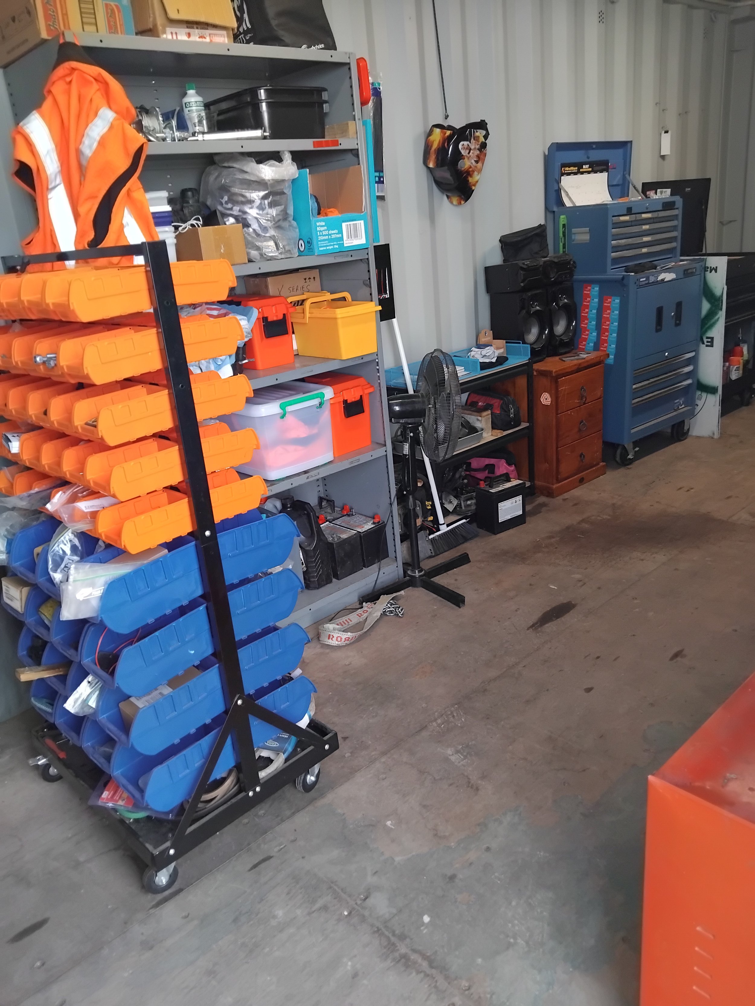 Storage area with shelves and toolboxes, an orange safety vest hanging on a shelf, a black and white portable fan, and a blue rolling tool cabinet.