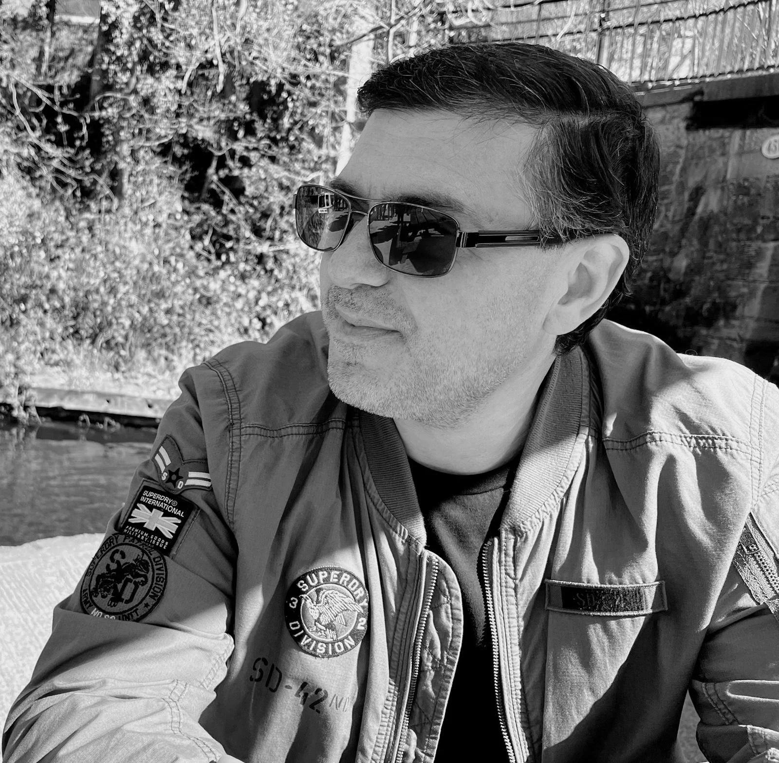 A man wearing sunglasses and a jacket with patches, sitting outdoors near a river and trees, with a fence in the background.