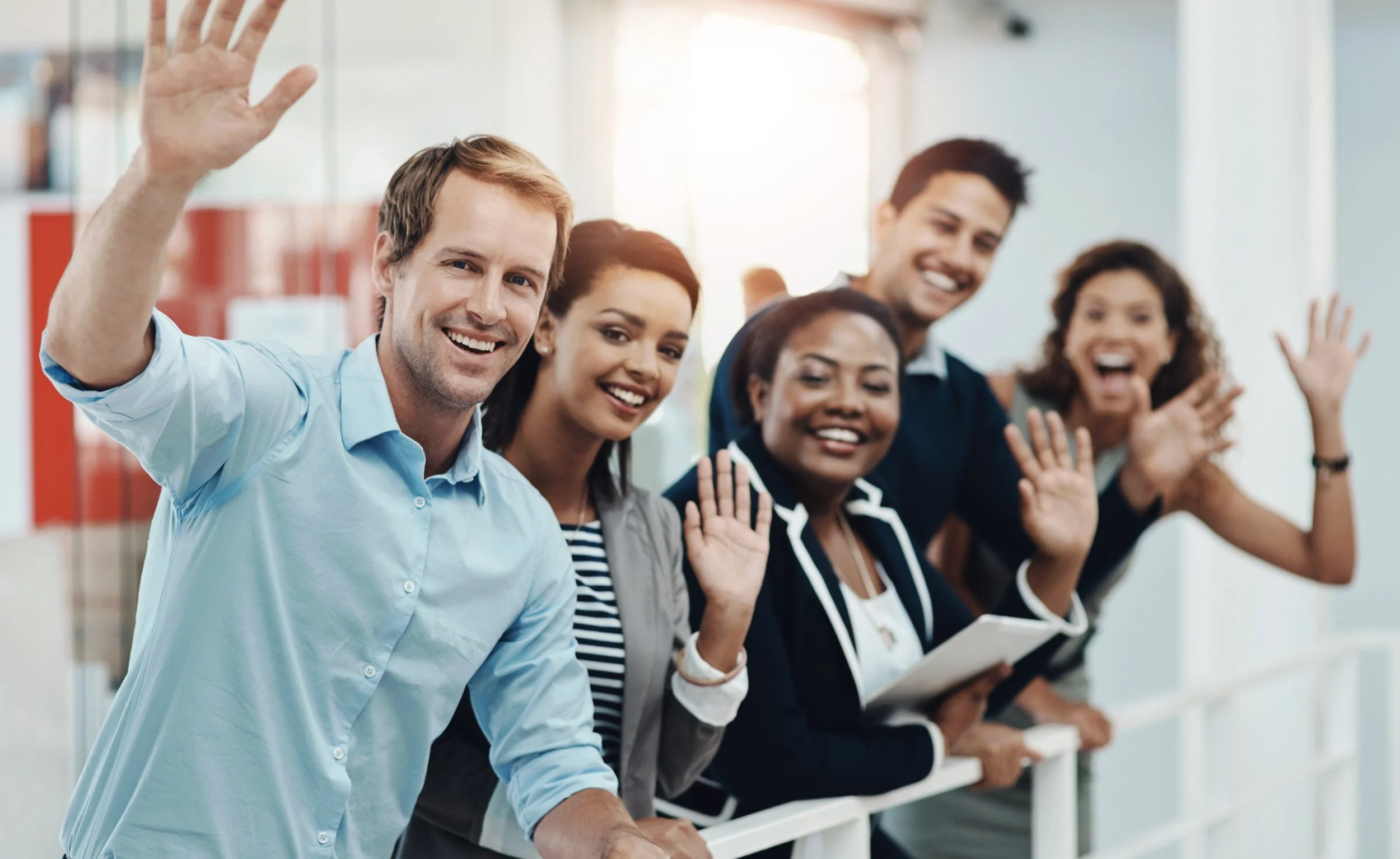 Group of five diverse people smiling and waving at an office or conference event.