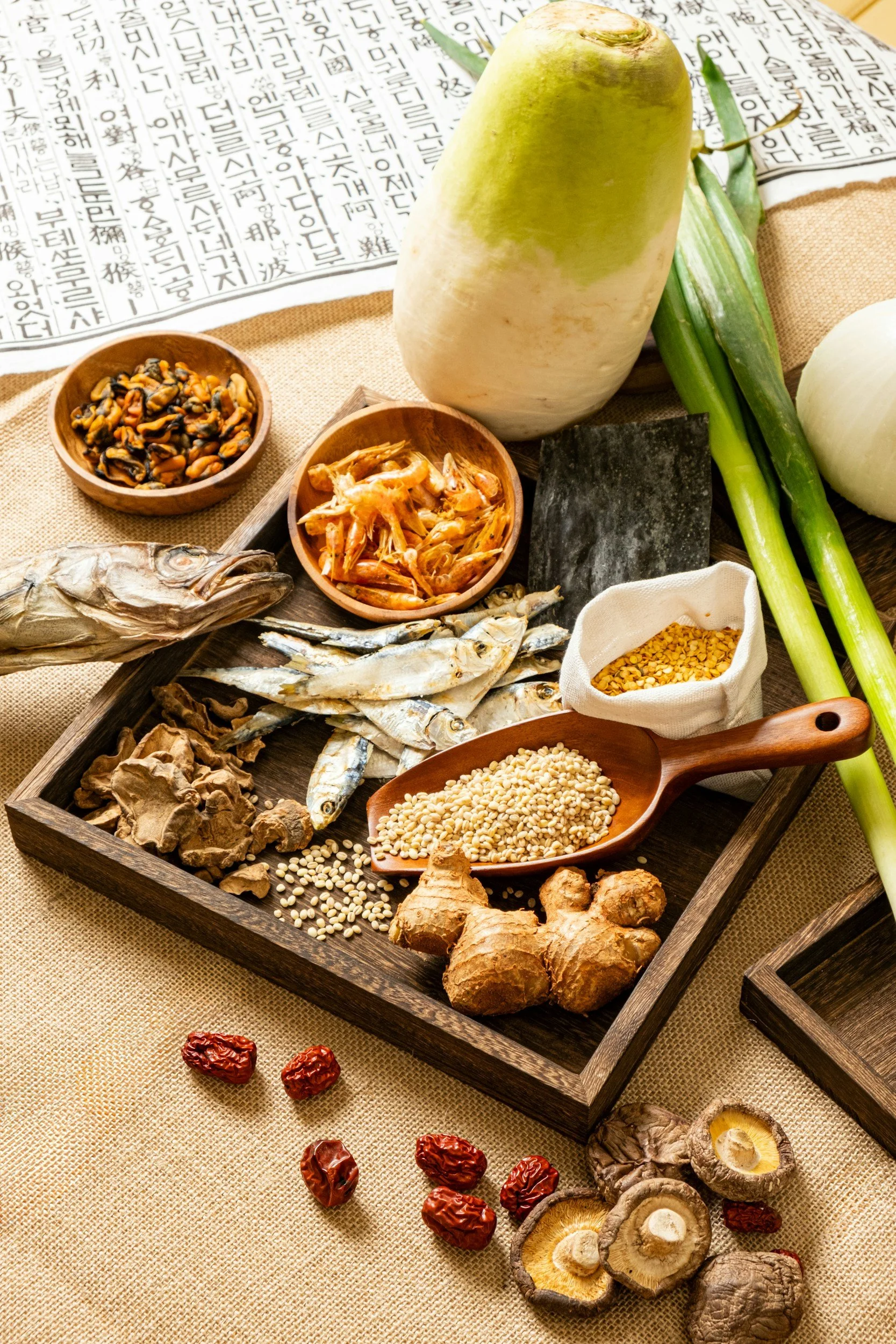 Traditional Korean herbal medicine ingredients arranged on a wooden tray, including dried roots, mushrooms, ginger, ginseng, dried dates, dried jujubes, black stone, sesame seeds, dried shrimp, and various herbs.
