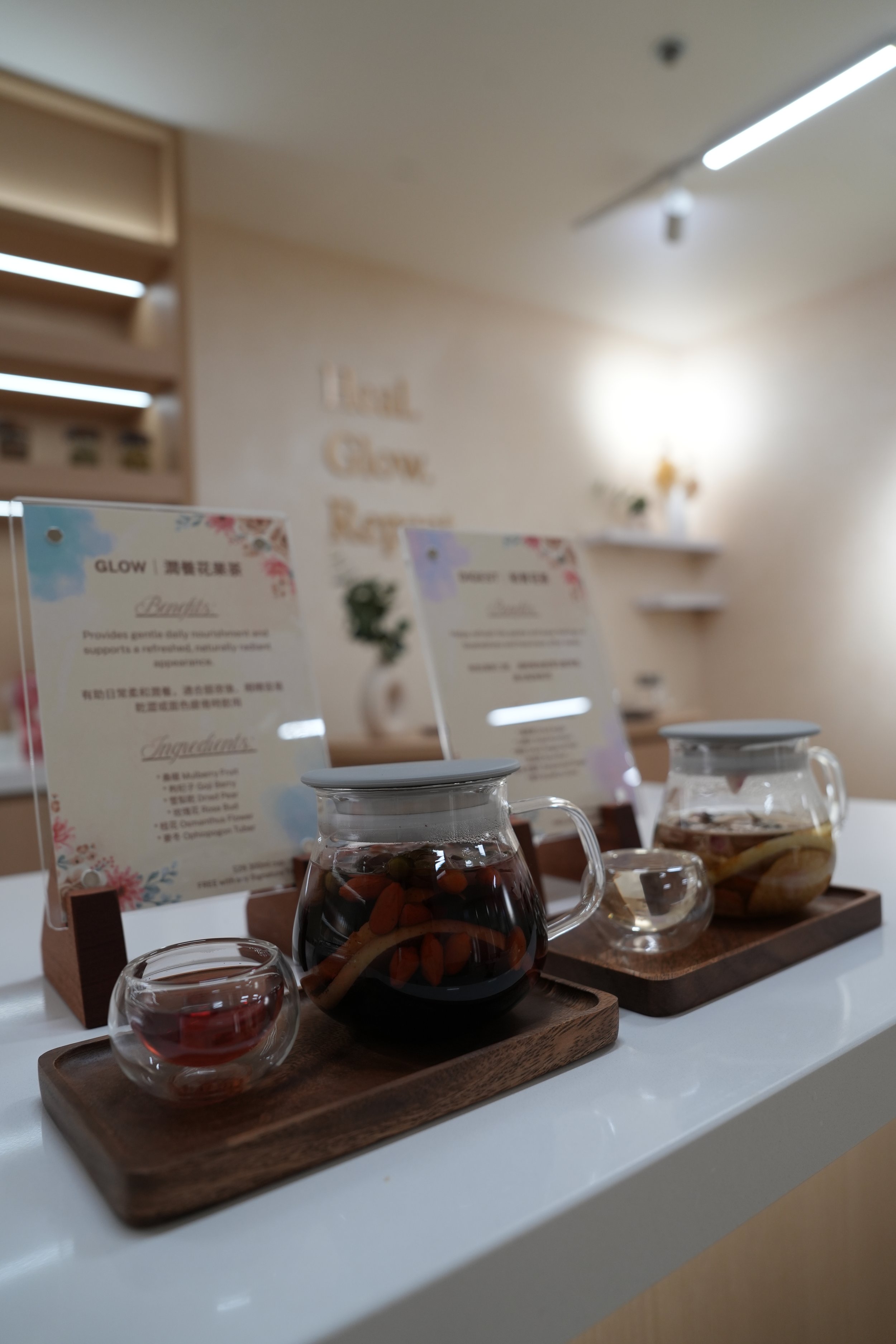 Three glass teapots with different herbal teas and wooden trays, with informational cards on a white table in a cafe or tea shop.