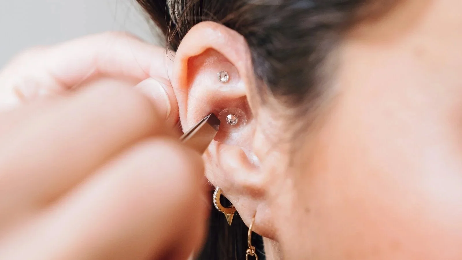 Close-up of a person's ear with multiple piercings, including two diamond studs, and a jewelry tool inserting or adjusting a jewelry piece in the lower piercing.