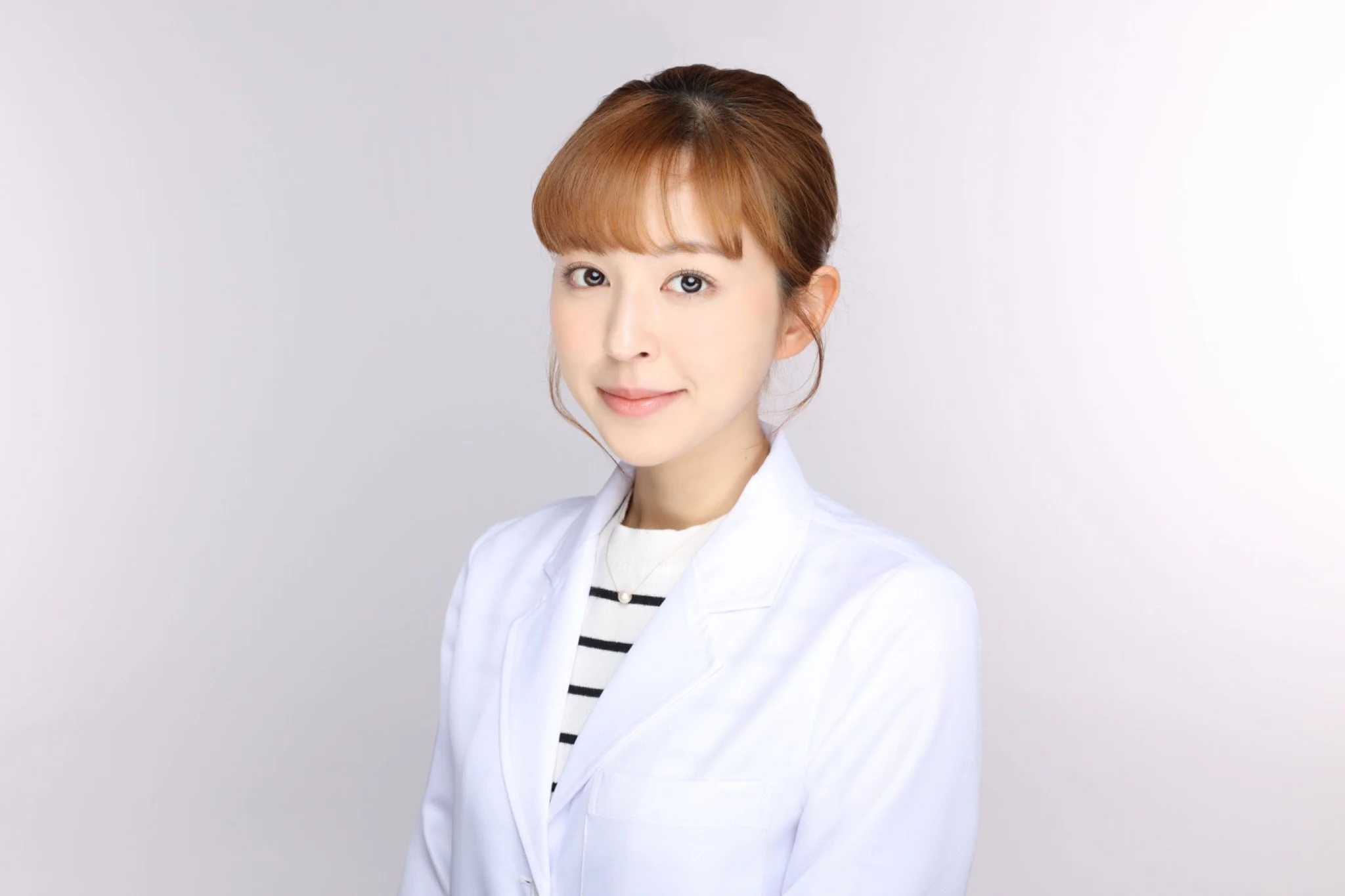 A woman with light skin and brown hair styled in a bun, wearing a white lab coat over a striped shirt, smiling softly against a plain light background.