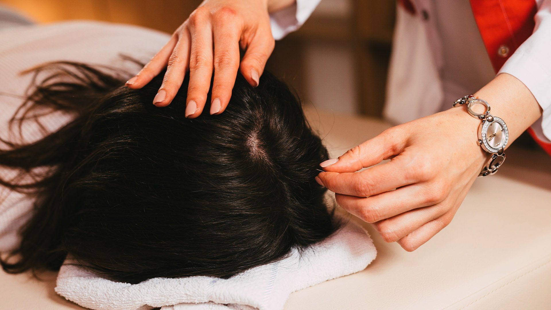 A person with light skin is giving a head massage to another person with dark hair who is lying face down on a towel.