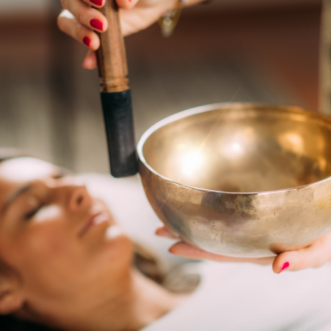 A woman lying down with eyes closed, receiving a sound healing session using a large metal singing bowl and a mallet, in a wellness or therapy setting.