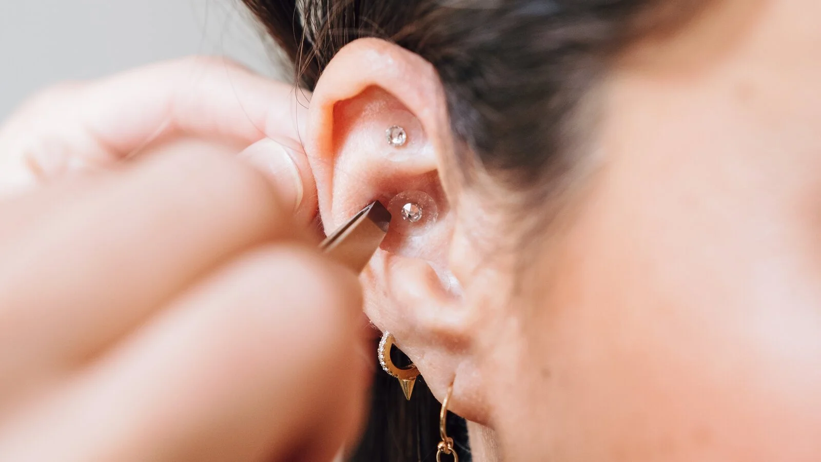 Close-up of a person having a piercing done on their earlobe, with several earrings already in place and a professional using a needle to insert a new earring.