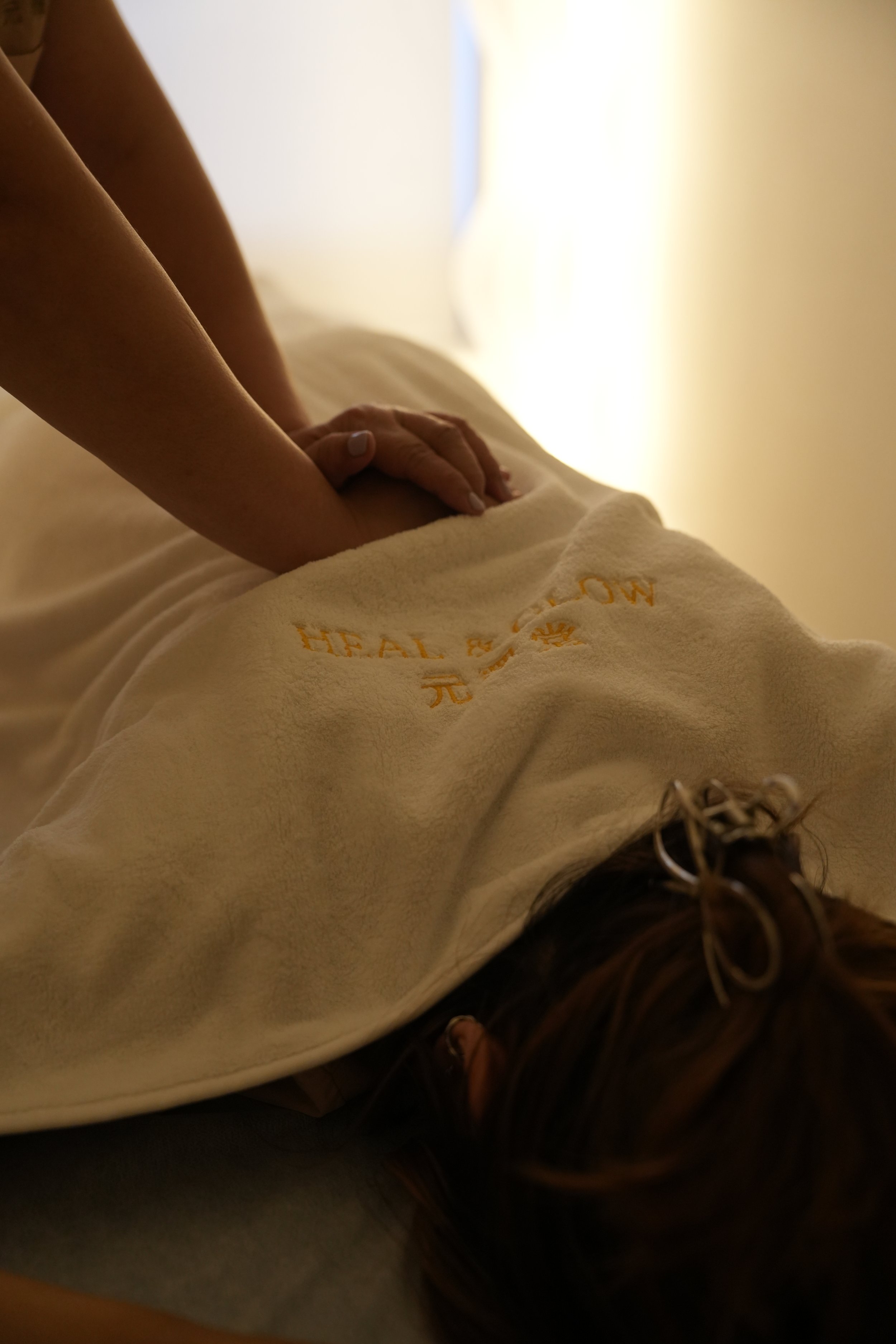Person lying down on a bed with hands resting on their abdomen, wearing a cream-colored shirt with orange text and laced shoes, hair tied with a hairband, in a softly lit room.