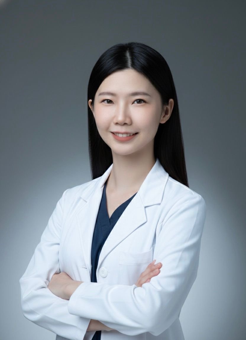 A young Asian woman with long black hair and a warm smile wearing a white medical lab coat and a dark scrub shirt underneath, standing with arms crossed against a plain gray background.