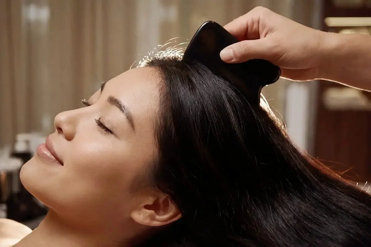 A woman with long black hair is getting her hair brushed by someone using a black hairbrush in a salon or spa setting.