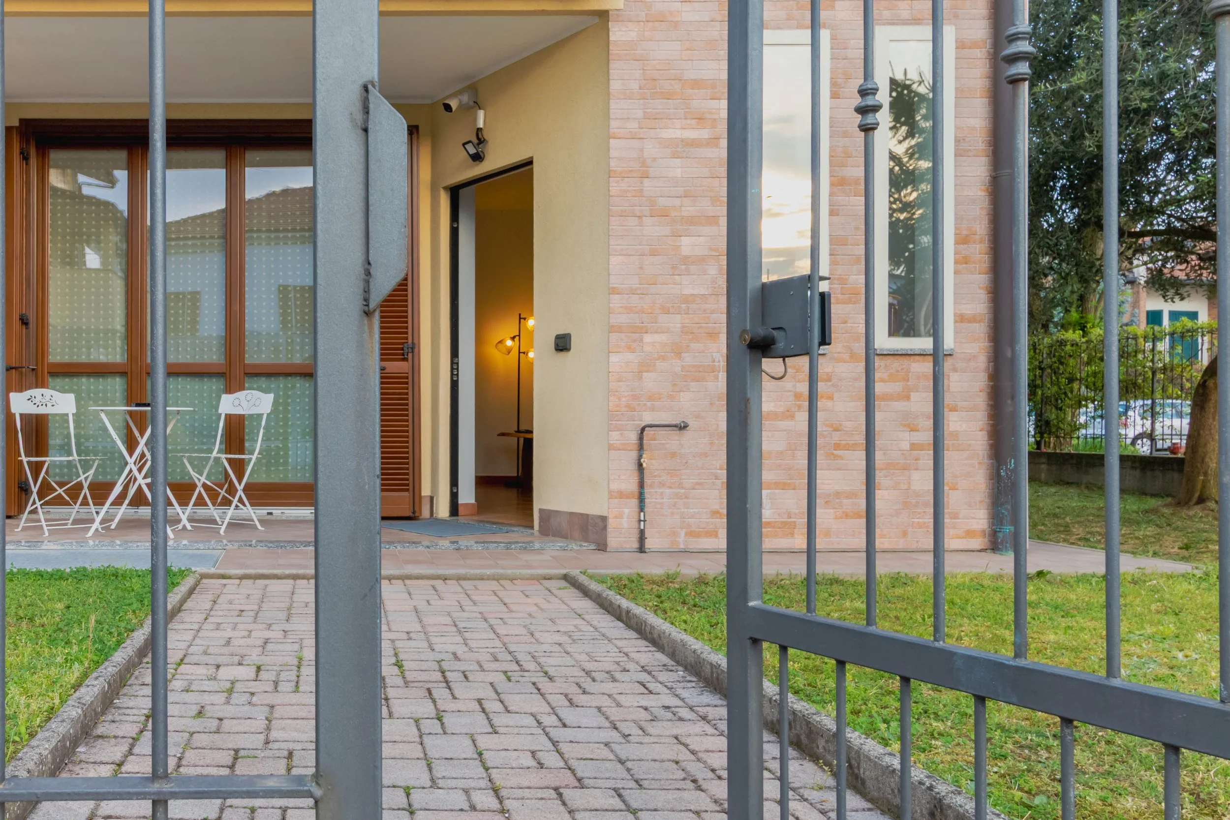 Veduta di un cancello in ferro che porta a una casa con giardino, con un'area esterna con tavolo e sedie, porte scorrevoli in vetro, pareti in mattoni e un portone aperto con una lampada
