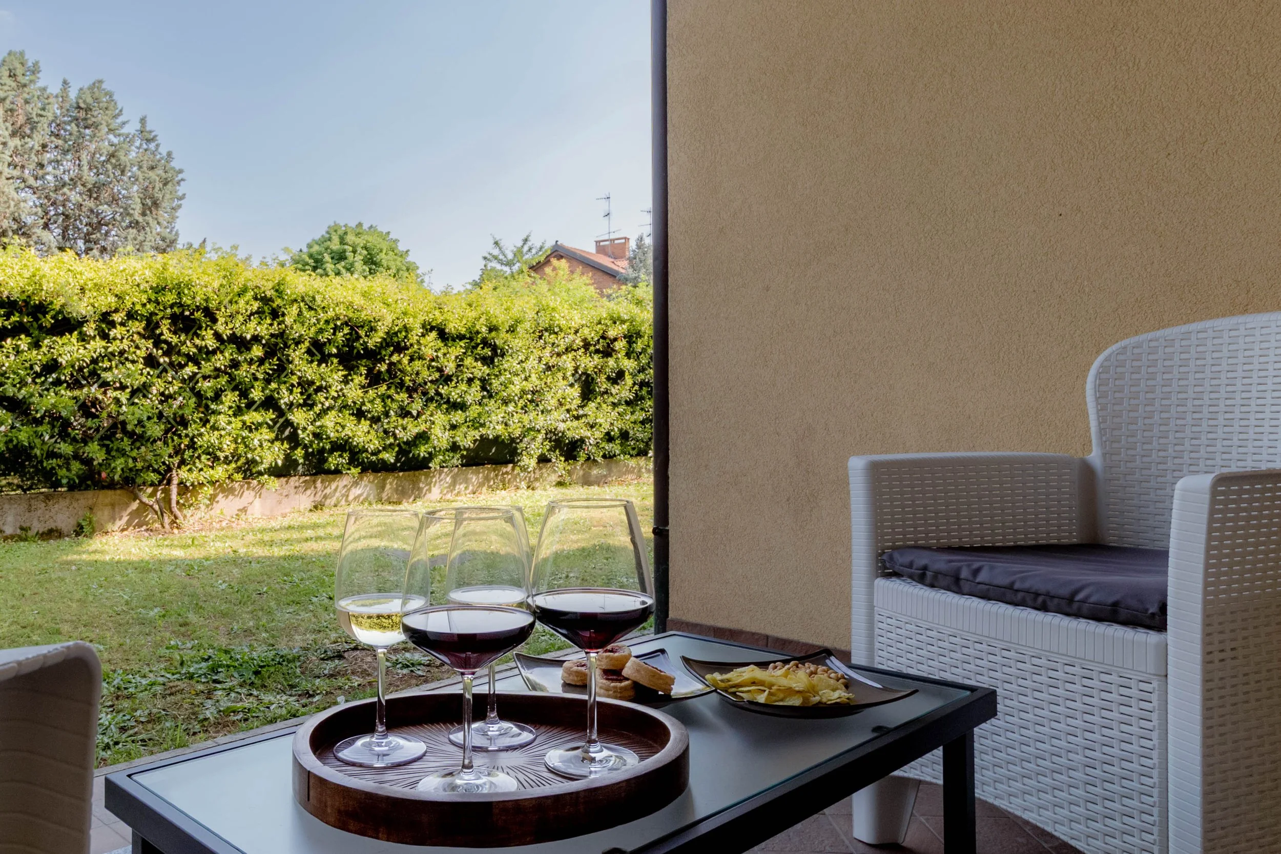 Vista di un patio con tavolo che ha quattro bicchieri di vino, un vassoio con tapas, formaggio, pane e altri snack. Sullo sfondo si vedono un giardino con cespugli verdi e alberi, e una casa con tetto rosso.