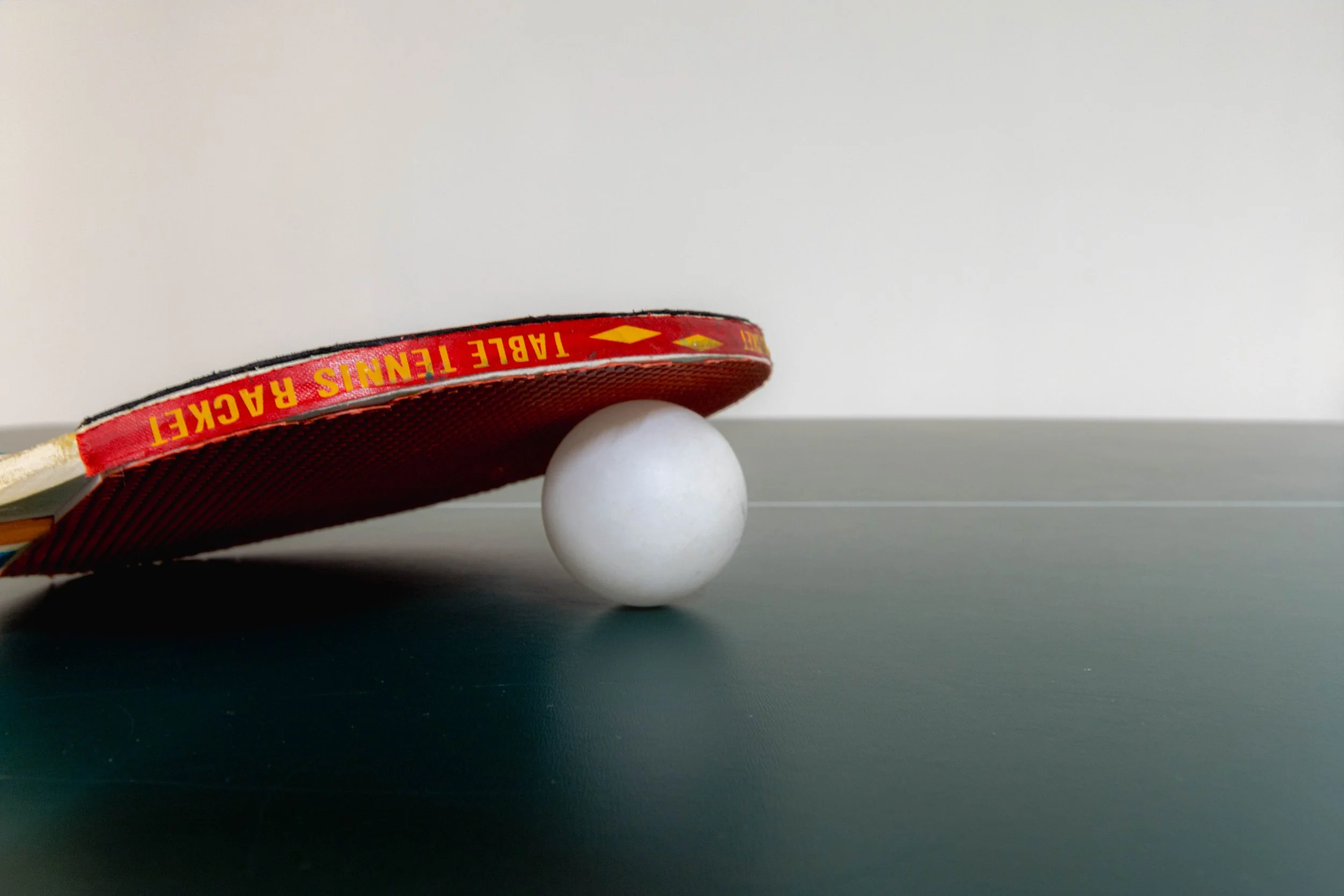 Una racchetta da ping pong rossa e nera appoggiata su due palline da ping pong una sopra l'altra, con uno sfondo neutro.