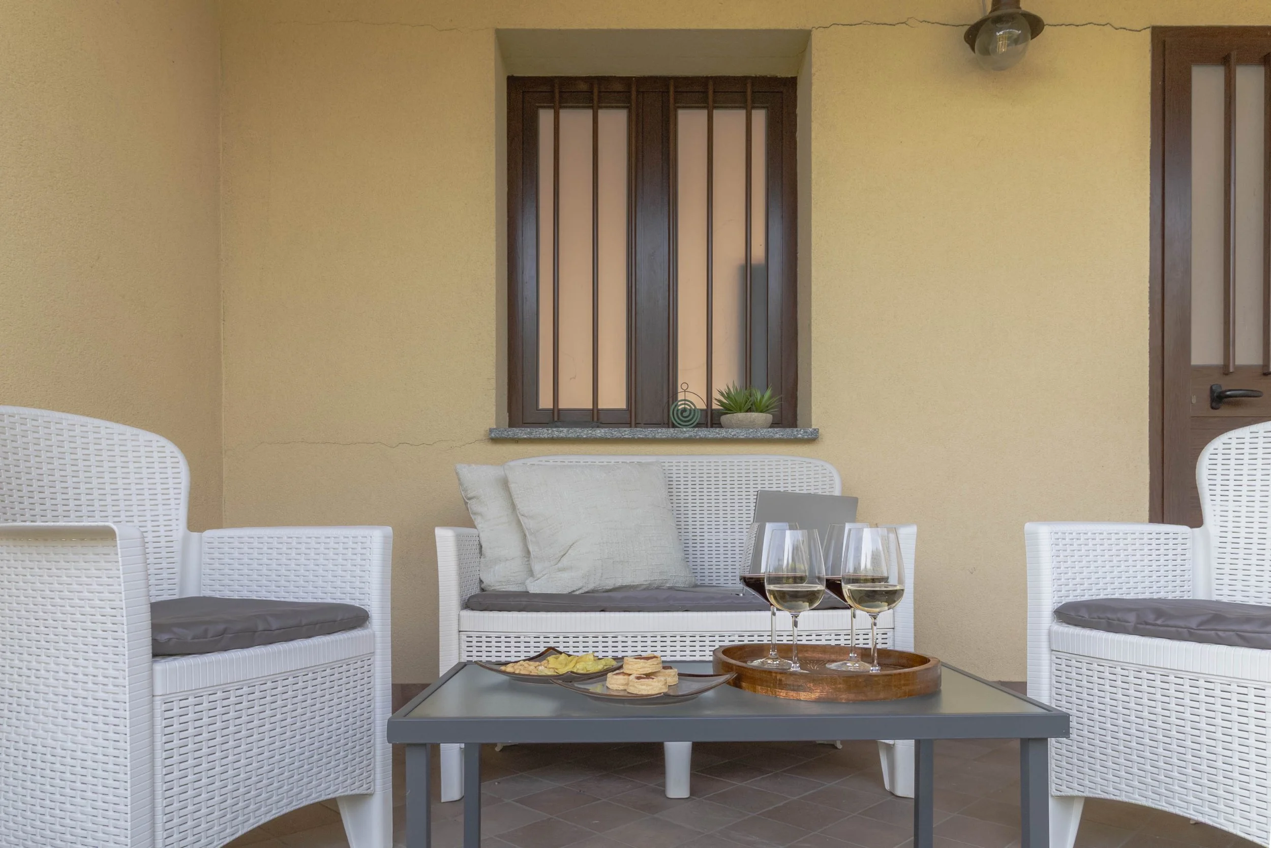 Zona esterna con pareti gialle, sedie bianche e un tavolo con acqua e snack. Sullo sfondo finestra con grate in ferro e porta in legno.