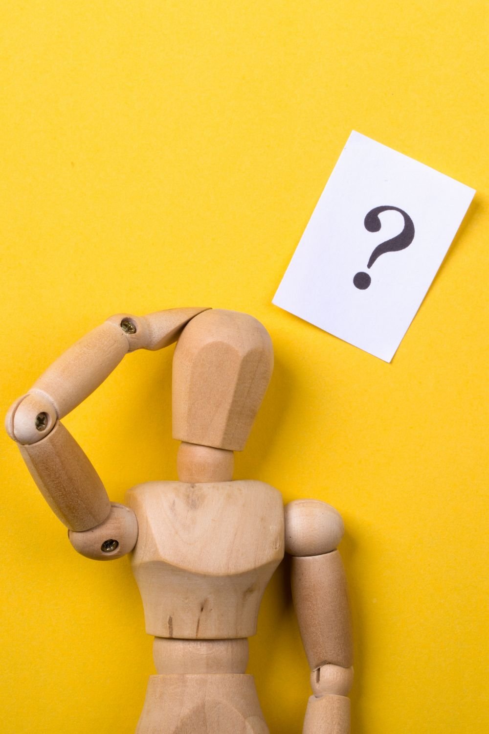 Una figura umana di legno, con la mano sulla testa, su uno sfondo giallo, con un cartello bianco con punto interrogativo.