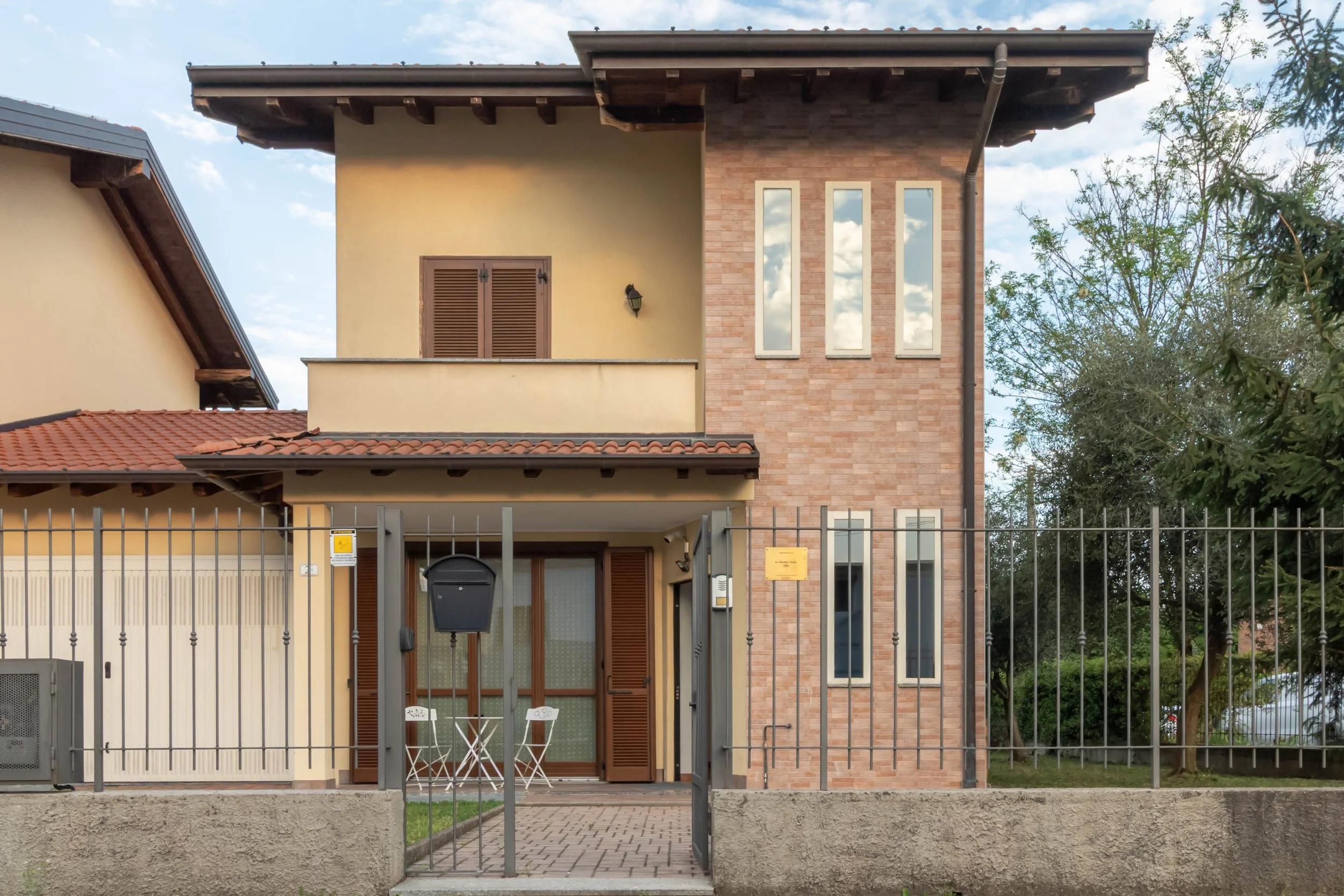 Casa moderna con facciata metà in mattoni e metà in stucco, finestrature alte e strette, terrazza con porta a vetri e sedie e tavolino bianchi, recinzione metallica, albero a destra, cielo nuvoloso.