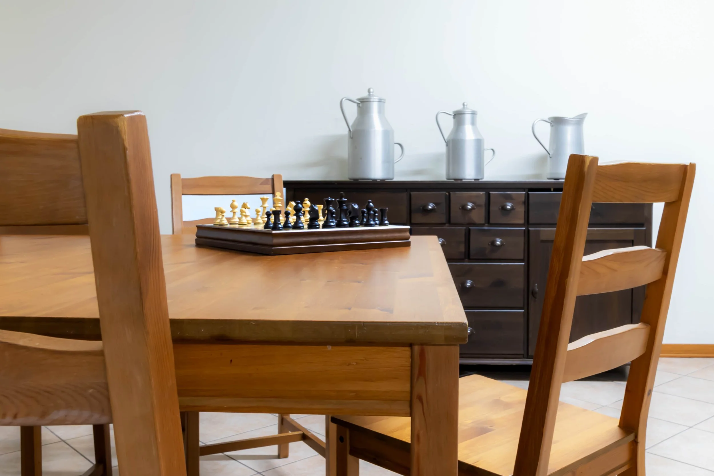 Tavolo di legno con scacchi e sedie di legno in una stanza con credenza scura e decorazioni di stince d'acciaio.