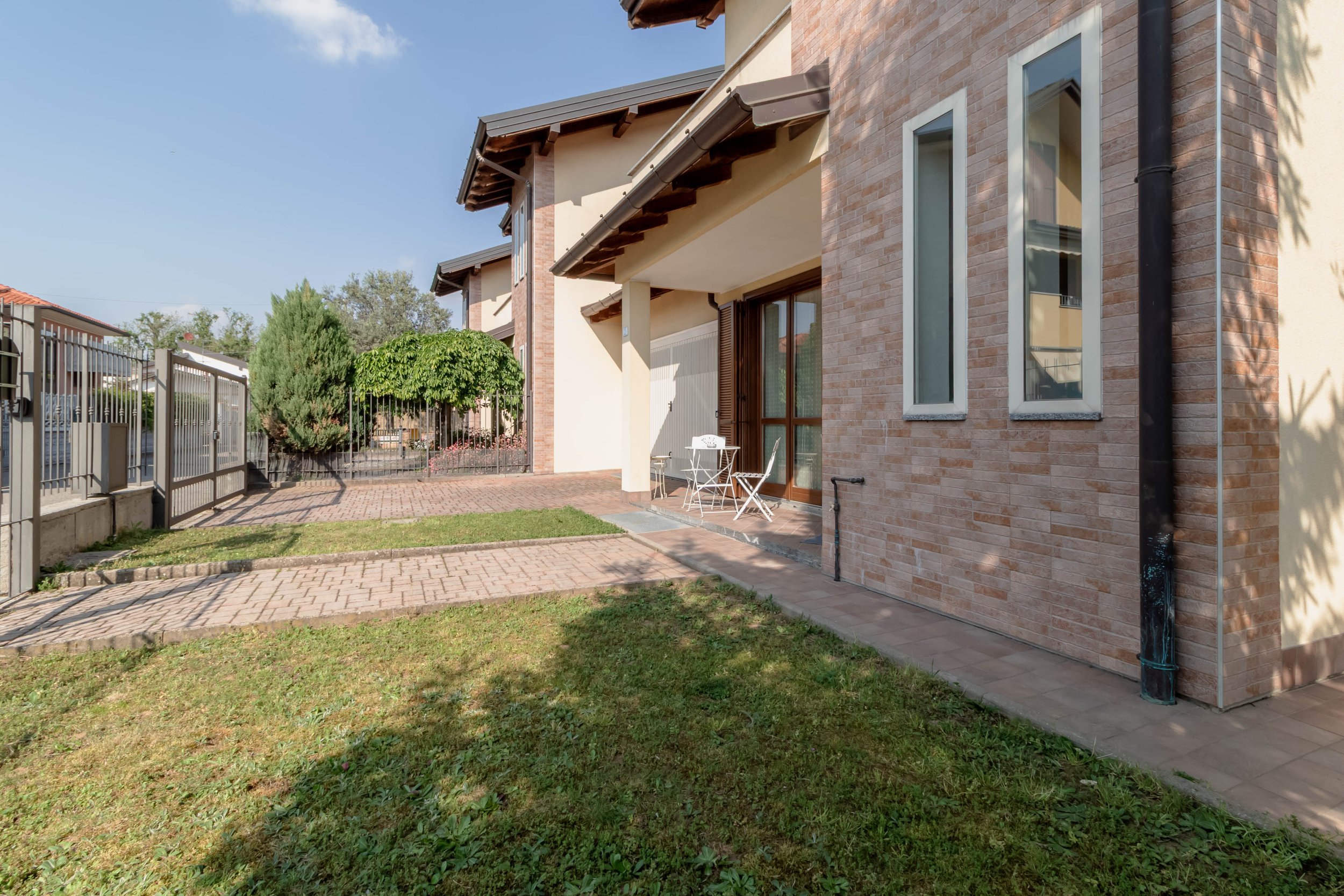 Esterno di una moderna casa con pareti in mattoni e grandi finestre, con un'area di giardino e un tavolo con sedie bianco sotto il balcone.
