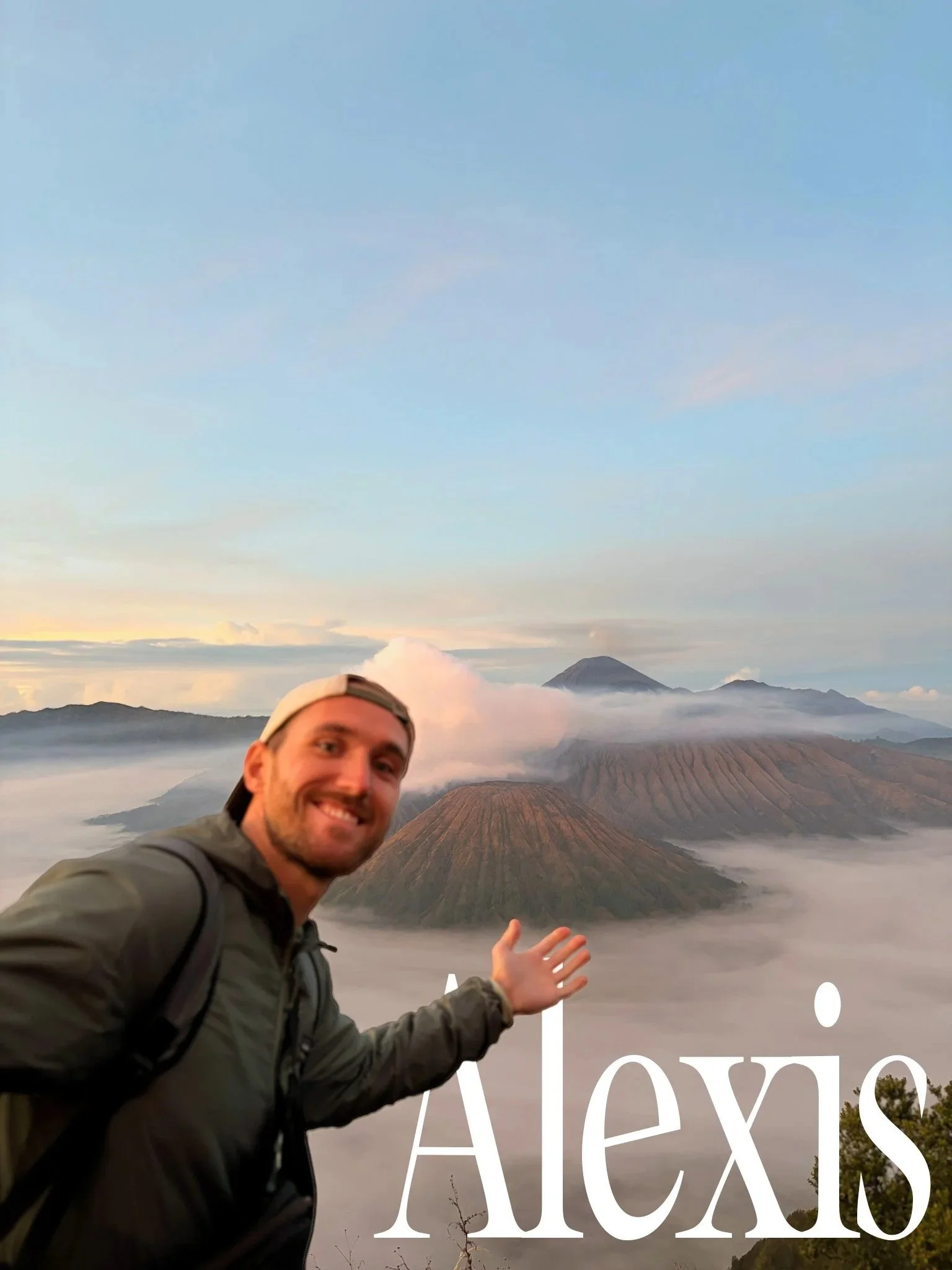 Un homme souriant, portant une veste noire, devant un paysage de volcans et de montagne avec un ciel partiellement nuageux, en arrière-plan, probablement lors d'une excursion ou d'une visite touristique.
