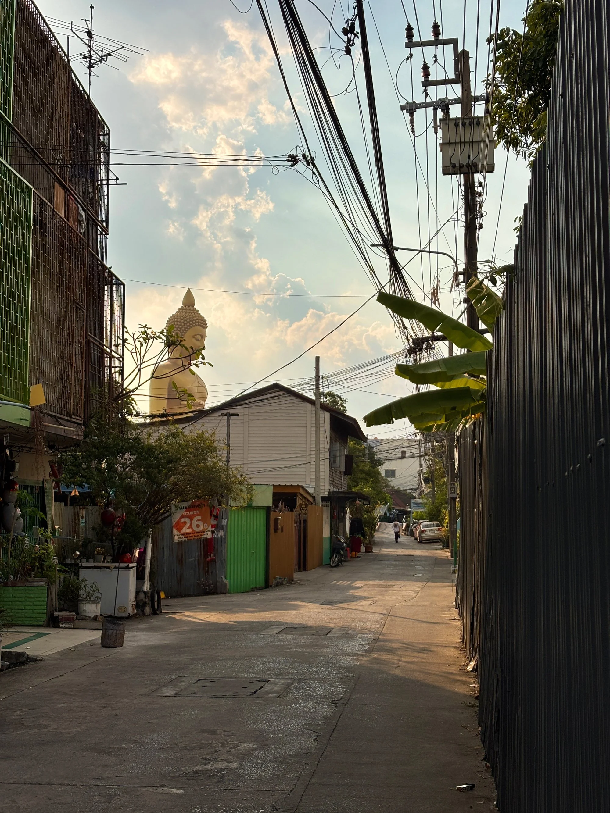 Une rue urbaine avec un temple bouddhiste en arrière-plan, des bâtiments en ferraille, des arbres et des câbles électriques contre un ciel partiellement nuageux.
