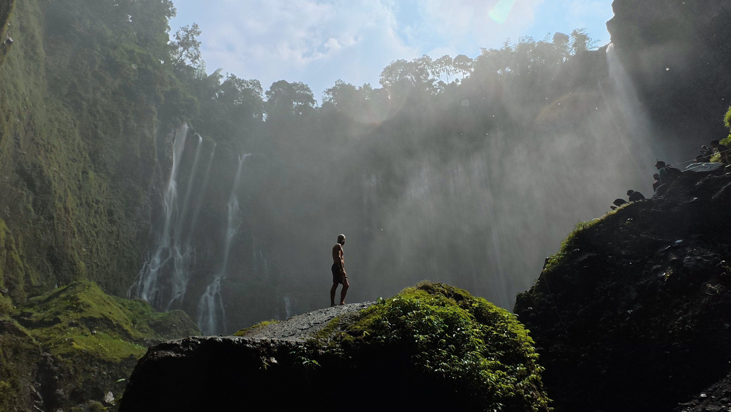 Personne debout sur un rocher devant une grande cascade entourée de végétation verte et de forêt, avec des rayons de soleil perçant le ciel au-dessus.