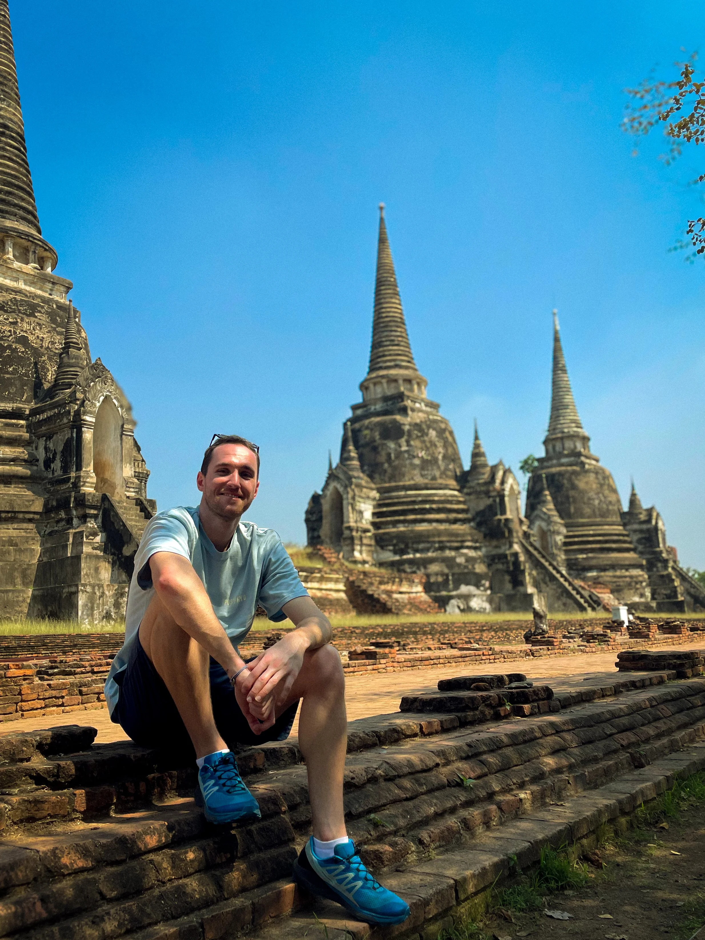 Un homme en vêtements de sport s'assoit sur les escaliers en pierre d'une ancienne pagode bouddhiste avec plusieurs tours pointues, sous un ciel bleu clair, dans un endroit historique en Thaïlande.