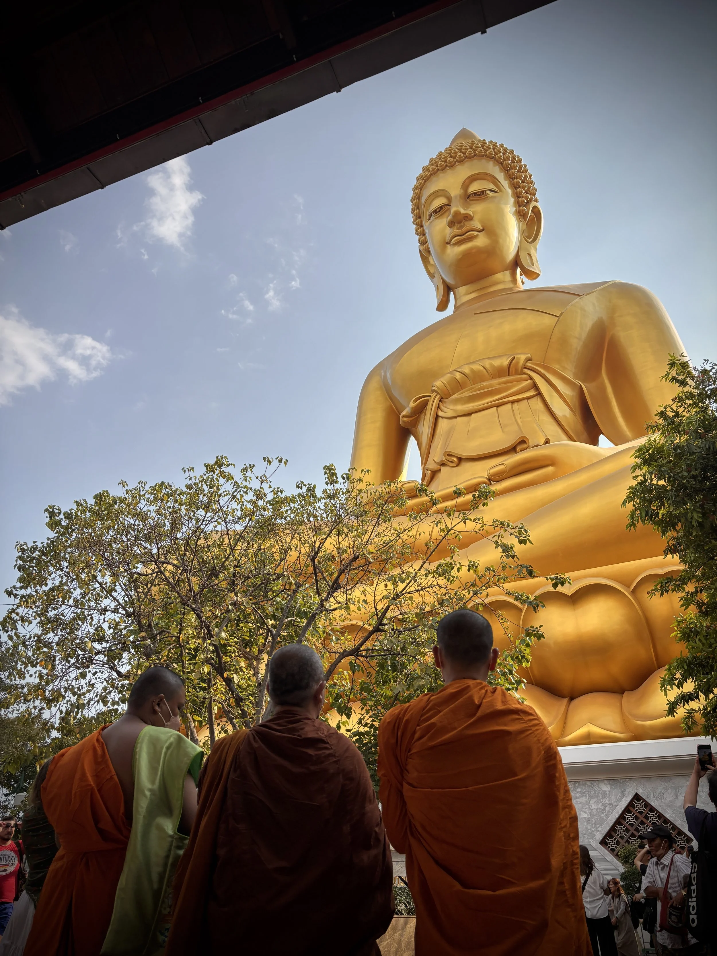 Un grand Bouddha doré en extérieur avec des personnes habillées en orange et en tenue décontractée autour, certains prenant des photos, arbres et ciel en arrière-plan.