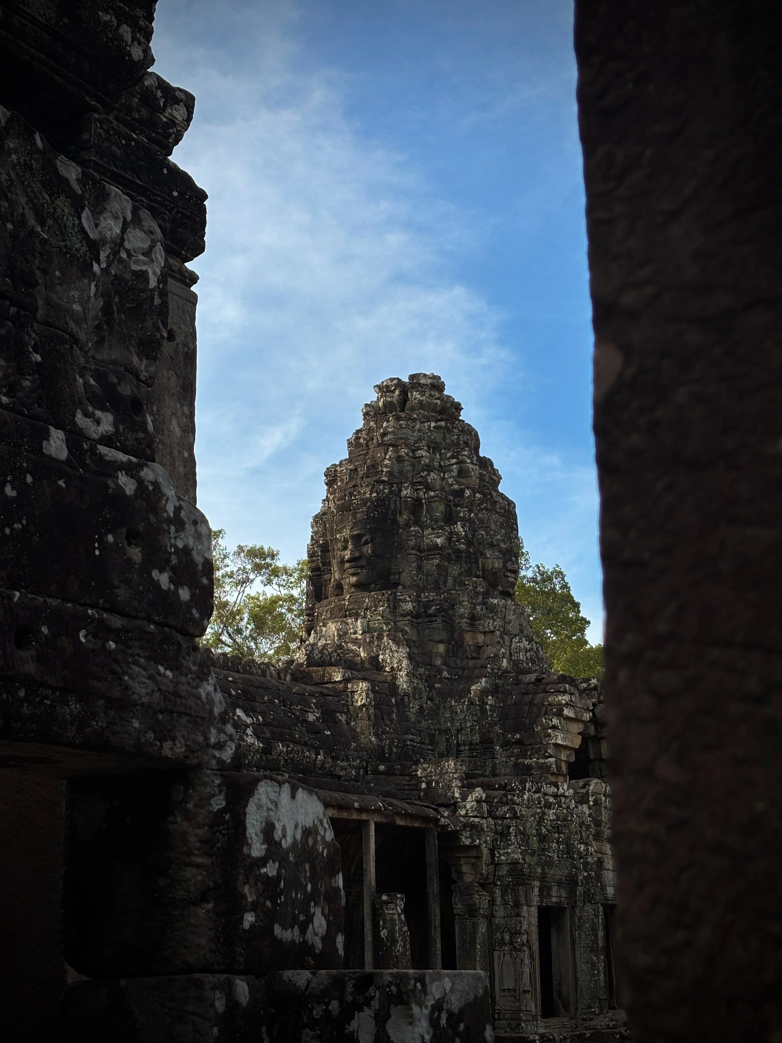 Vue d'une ancienne structure en pierre avec une grande tête sculptée, probablement d'une statue ou d'un temple, encadrée par des murs en pierre usés, sous un ciel bleu avec quelques nuages, dans un environnement naturel forêt.