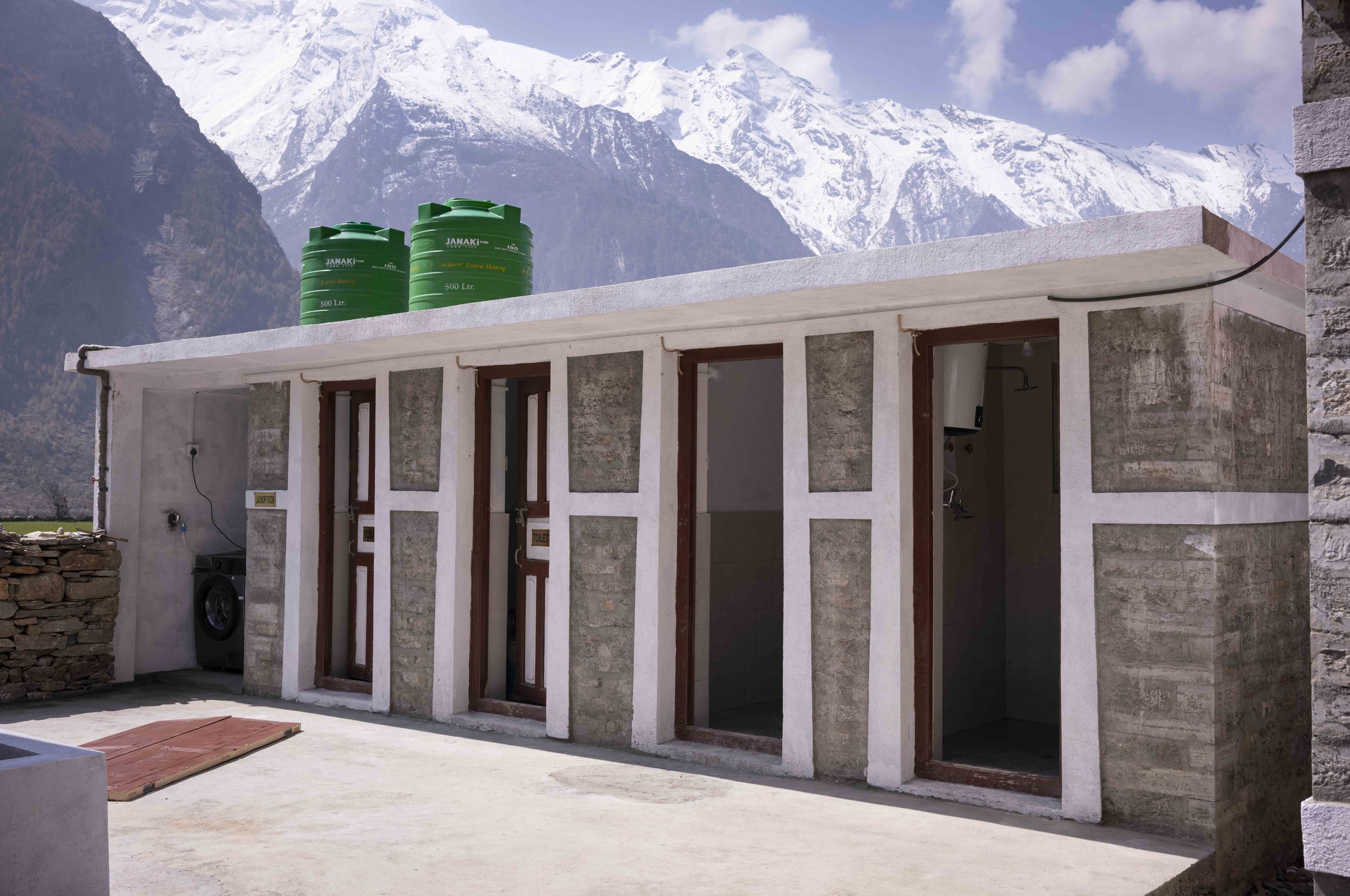 Building with multiple doorways, laundry area, and two green water tanks on the roof, set against snowy mountain peaks in the background.