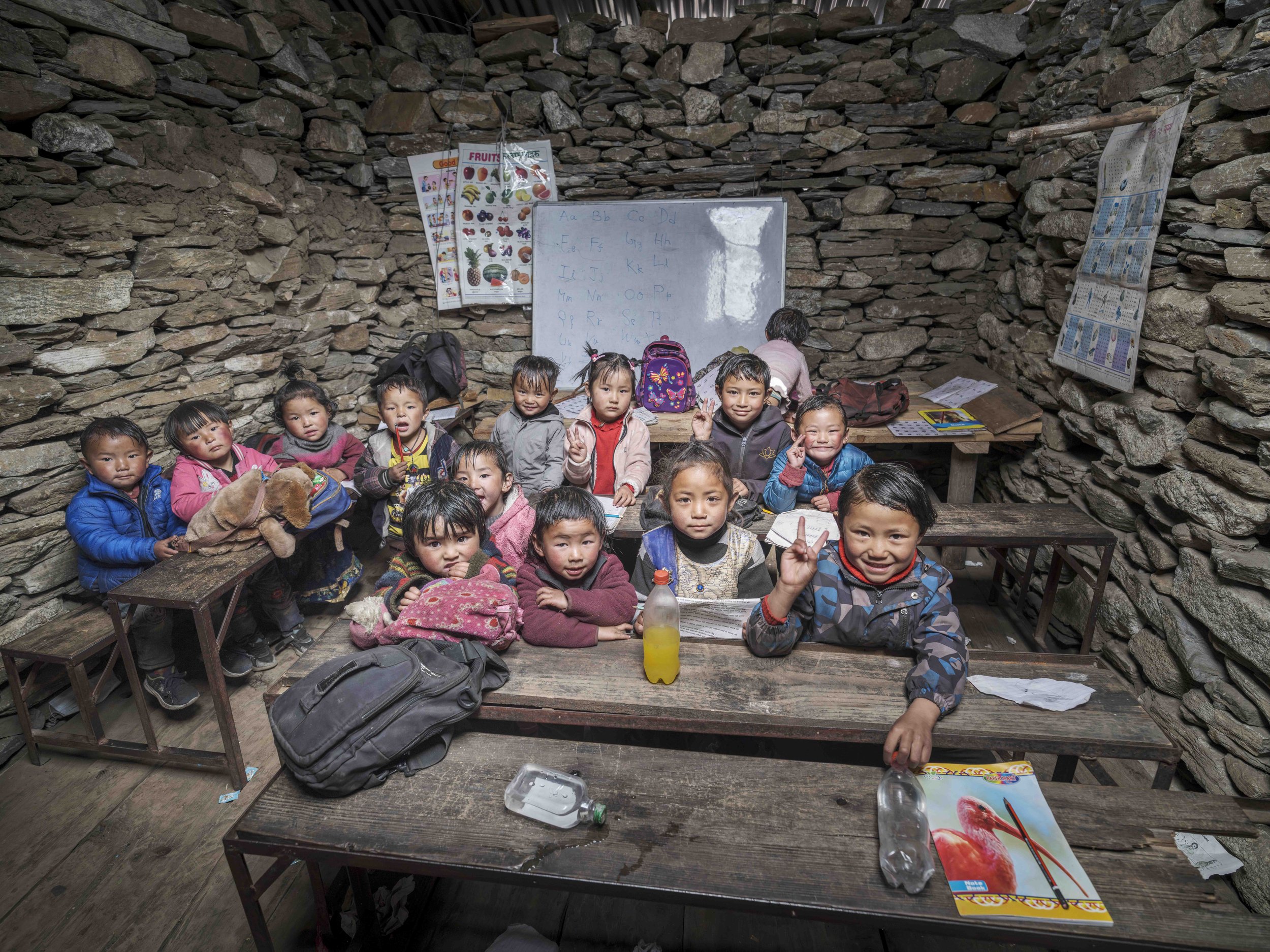 A group of children sitting at wooden tables inside a stone-walled classroom, with educational posters on the walls and various backpacks, notebooks, and bottled drinks on the tables.