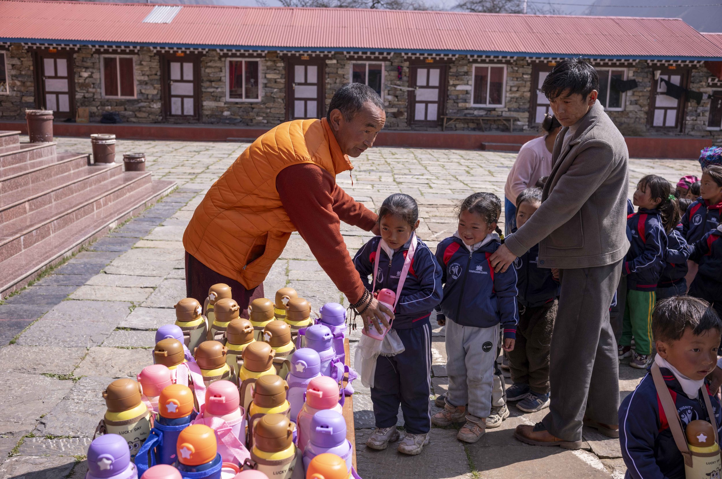 Children in school uniforms line up outdoors, with an adult woman and man handing out colorful water bottles during daytime.