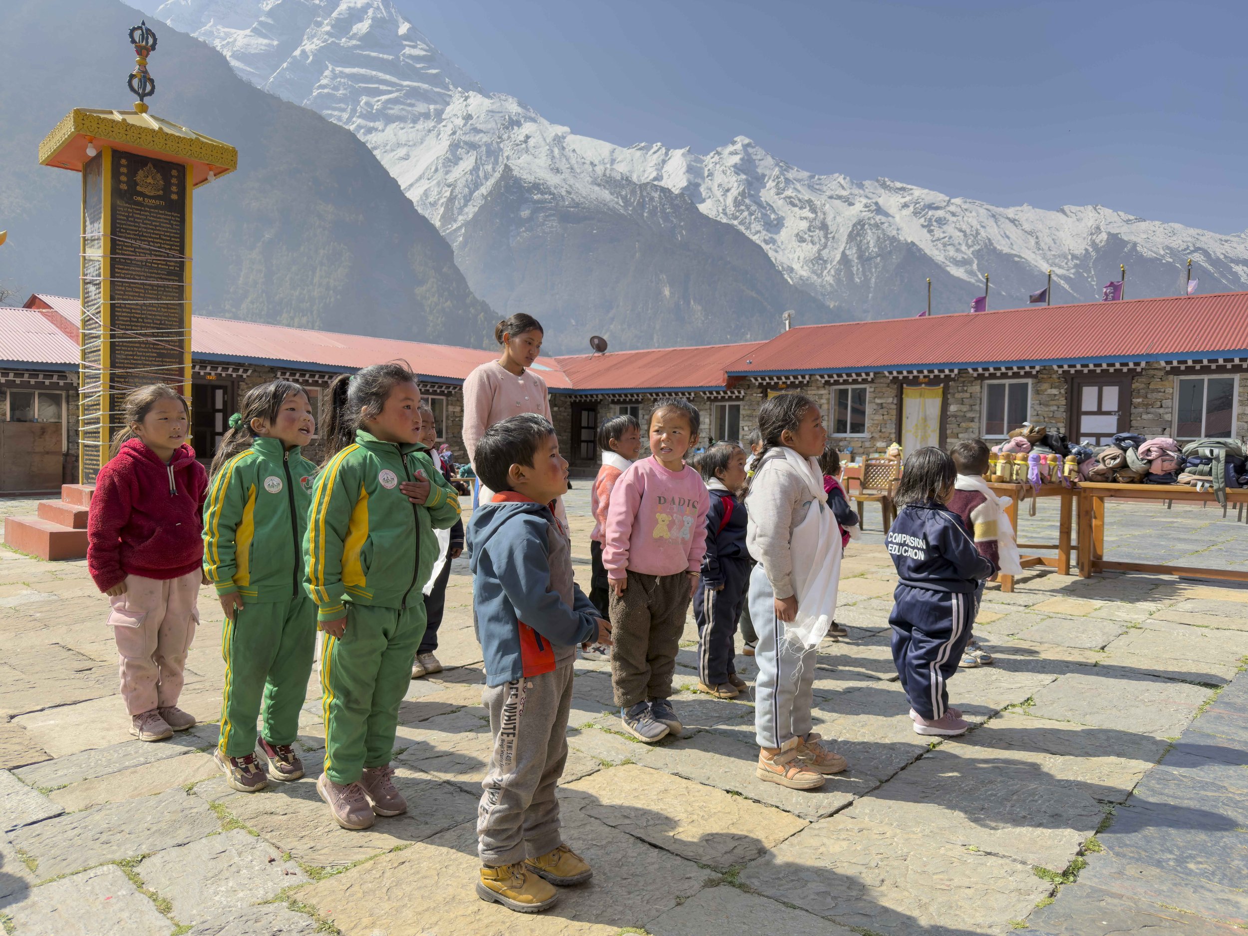 A group of children and a woman standing outdoors in front of a mountain range with snow-capped peaks, red-roofed buildings, and a clear blue sky.
