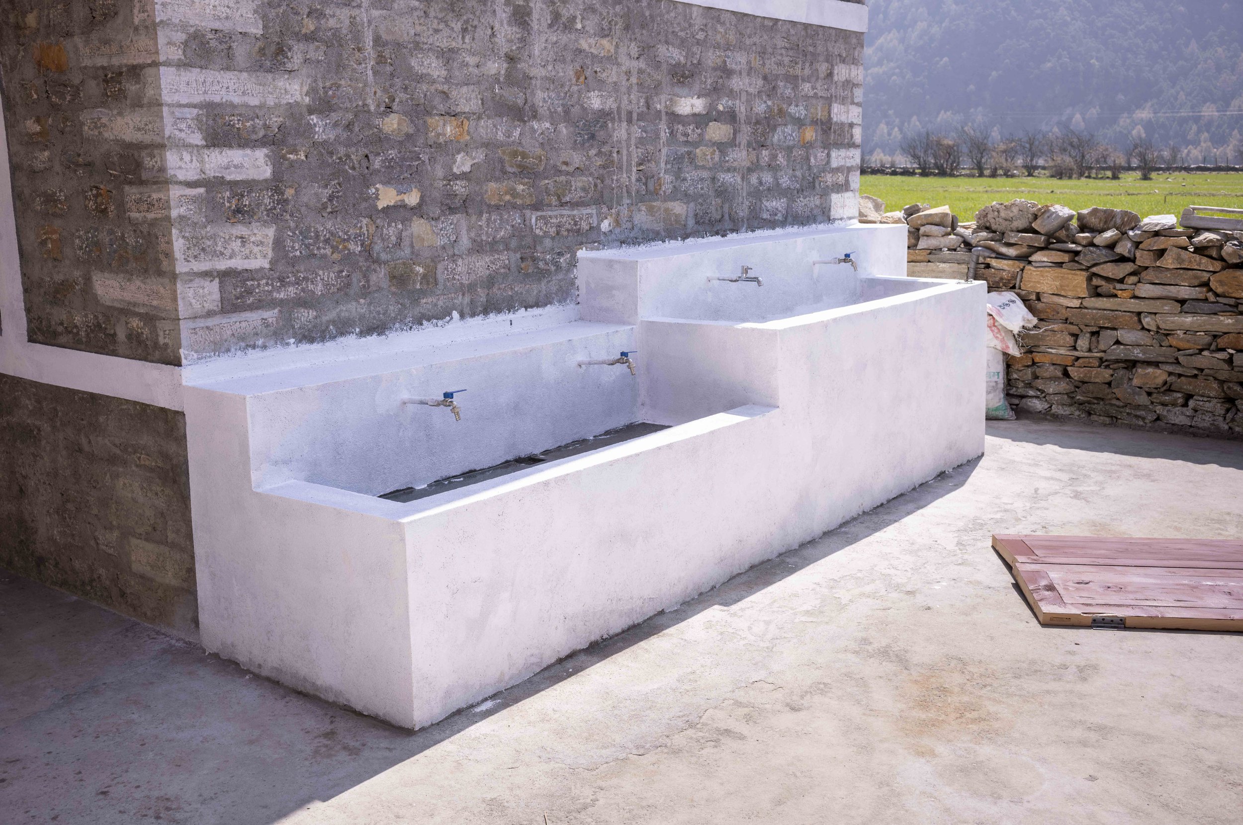 Outdoor scene featuring a stone wall with a white concrete sink with three faucets, a stone wall to the right, a grassy field, and trees in the background.