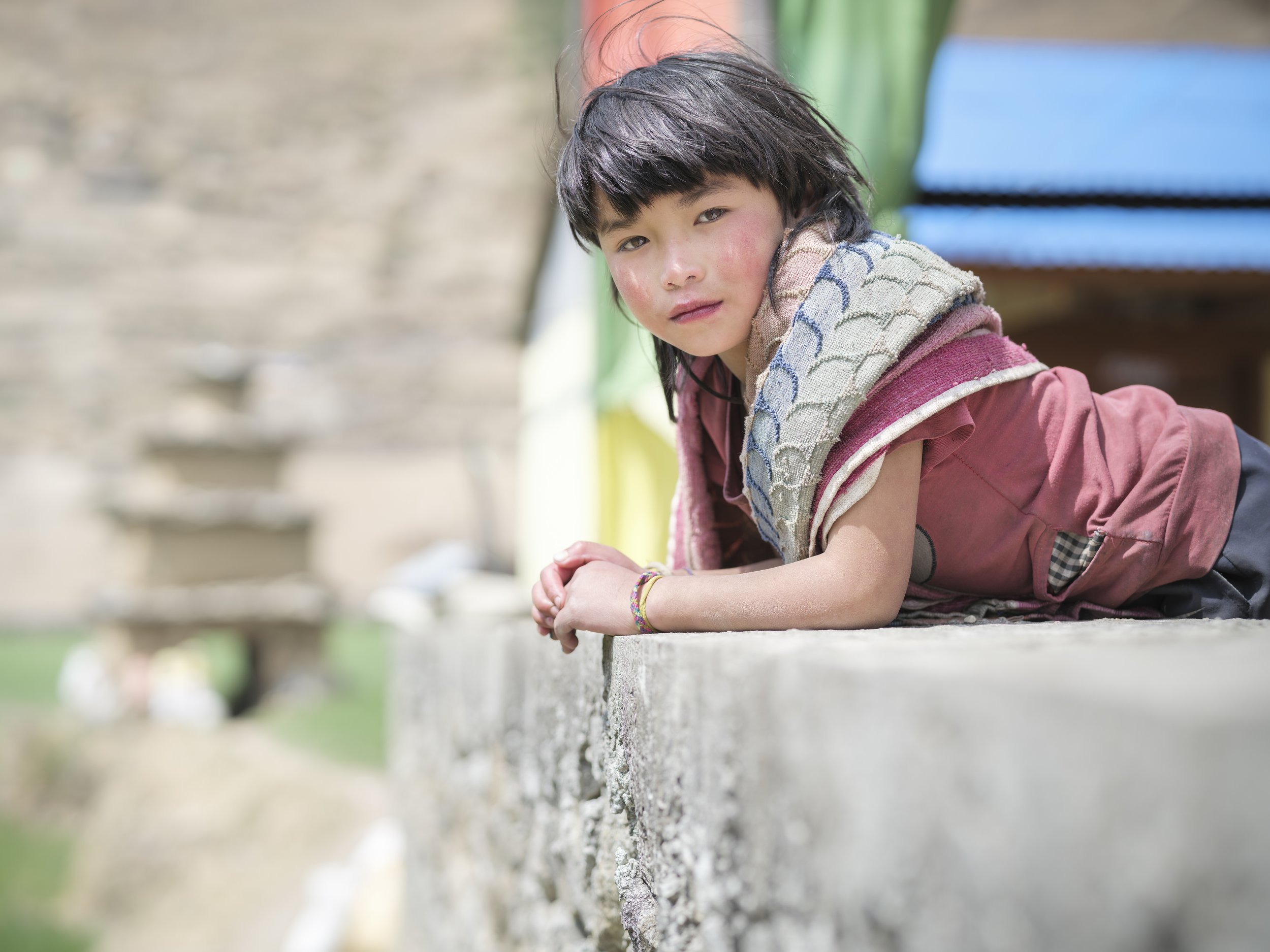 A young girl with dark hair leaning on a concrete ledge outdoors, wearing a pink jacket and a multicolored scarf, with a slightly serious expression.