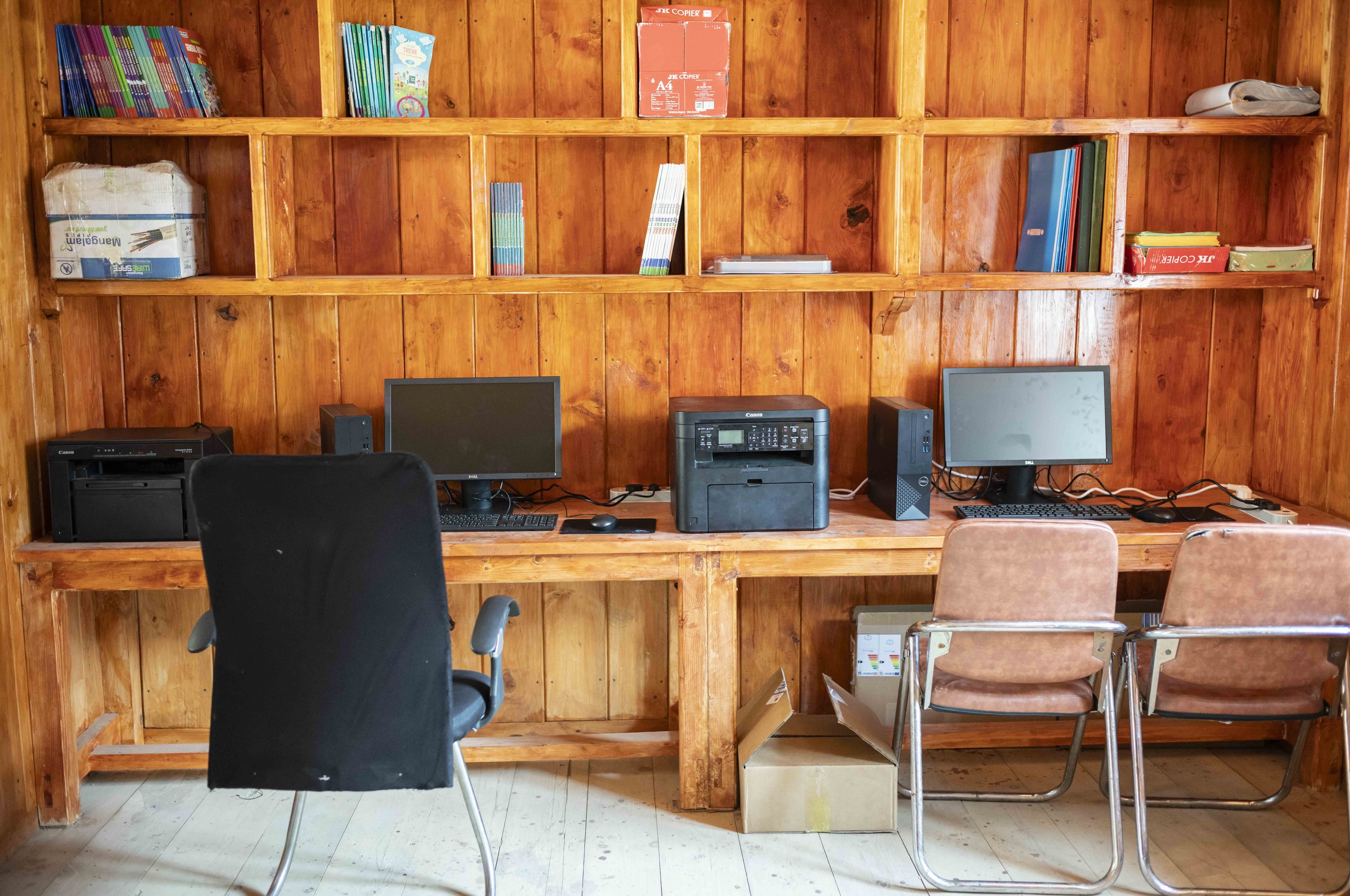A wooden office desk with two computer monitors, a printer, and some chairs and office supplies in front of a wooden wall shelf with books, binders, and paper stacks.