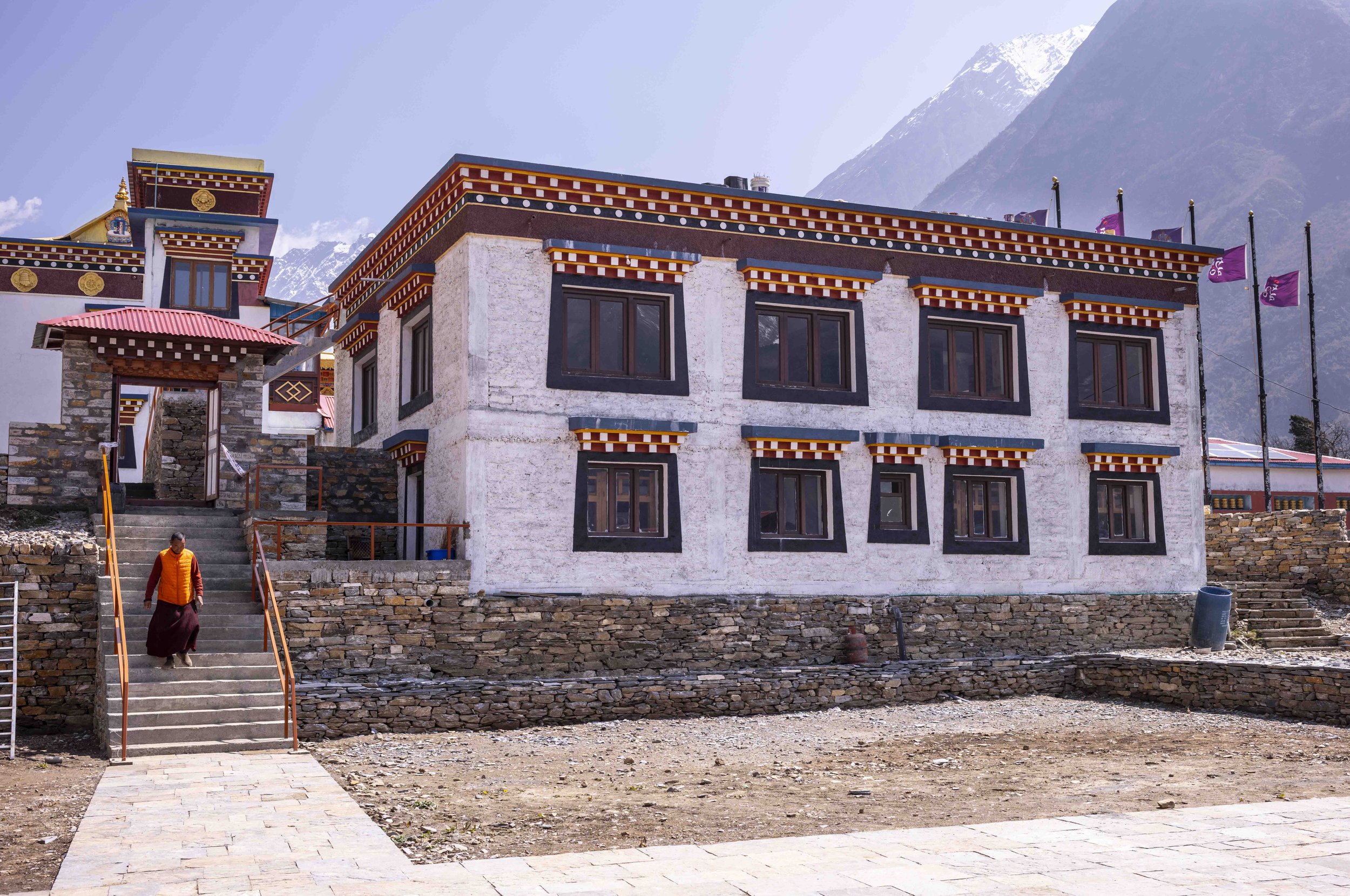 A traditional Buddhist monastery building with intricate colorful wooden eaves and windows, situated in a mountainous region with snow-capped peaks in the background. A person in warm clothing is walking up the stone steps leading to the entrance.