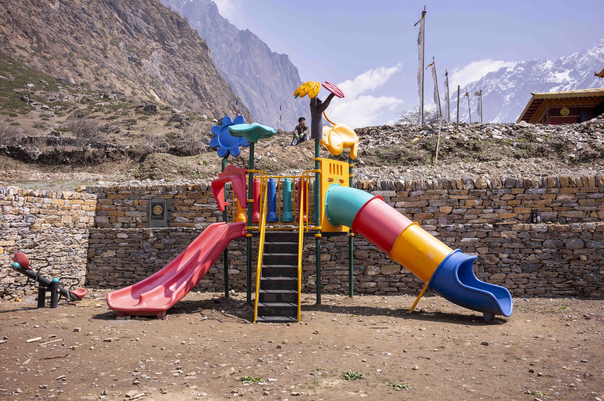 Colorful playground set with slides and climbing structures in a mountainous outdoor area with stone wall, flags, and snow-capped mountains in the background.