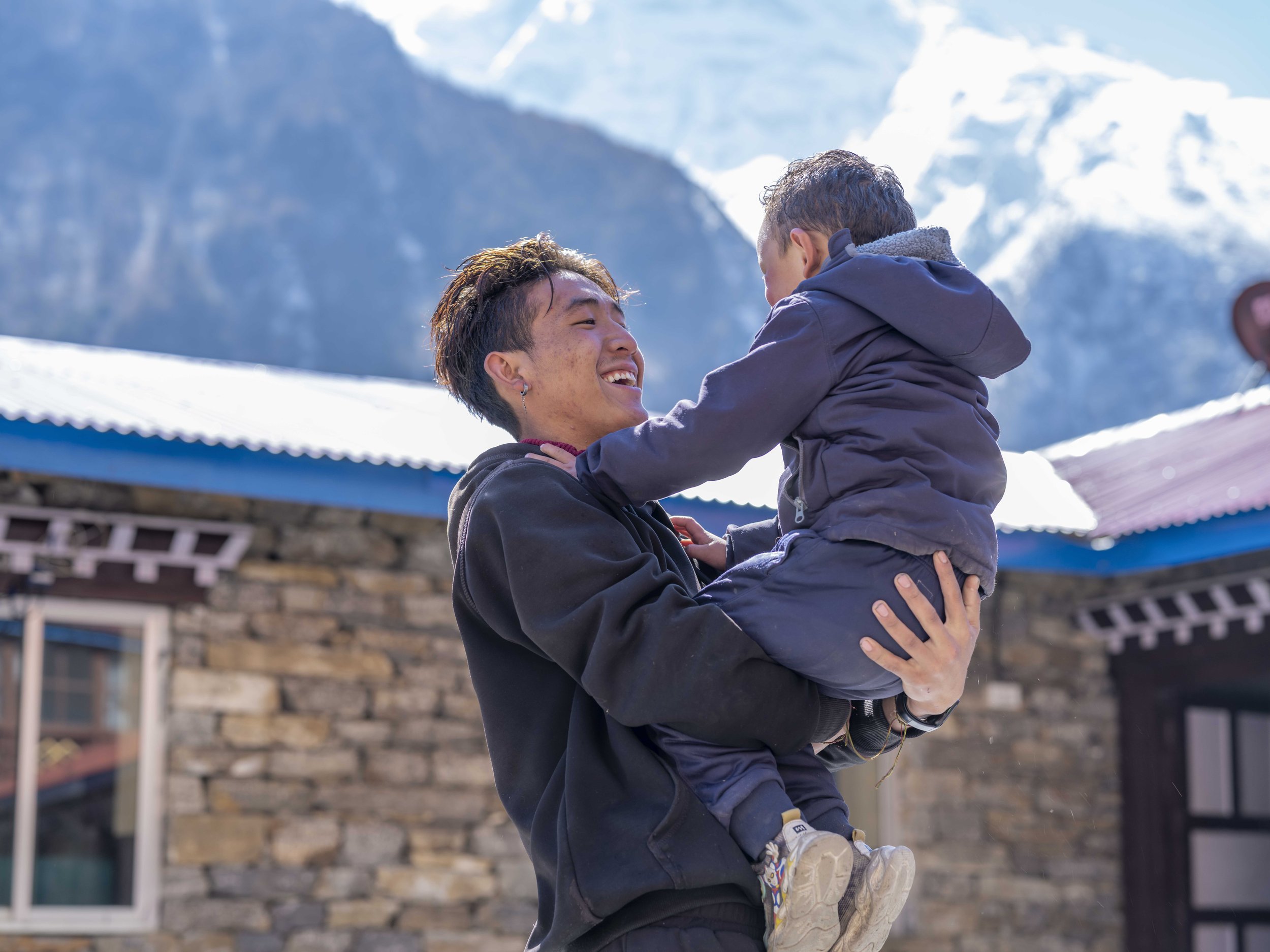 A person with short dark hair and earrings lifting a young boy in a winter coat and sneakers, with snow-covered mountains in the background.