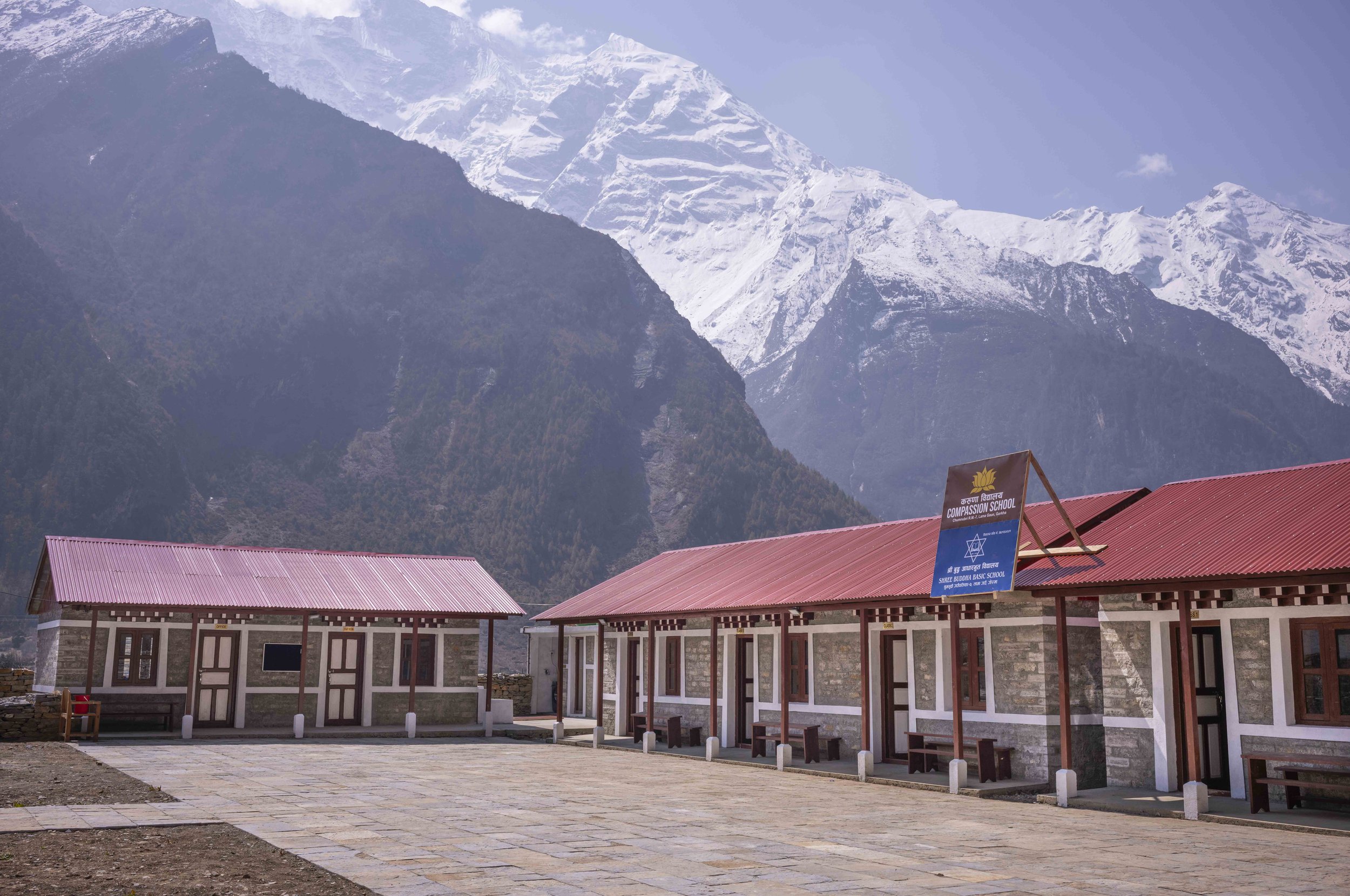 School buildings with red roofs situated in a mountainous region, with snow-capped peaks in the background.
