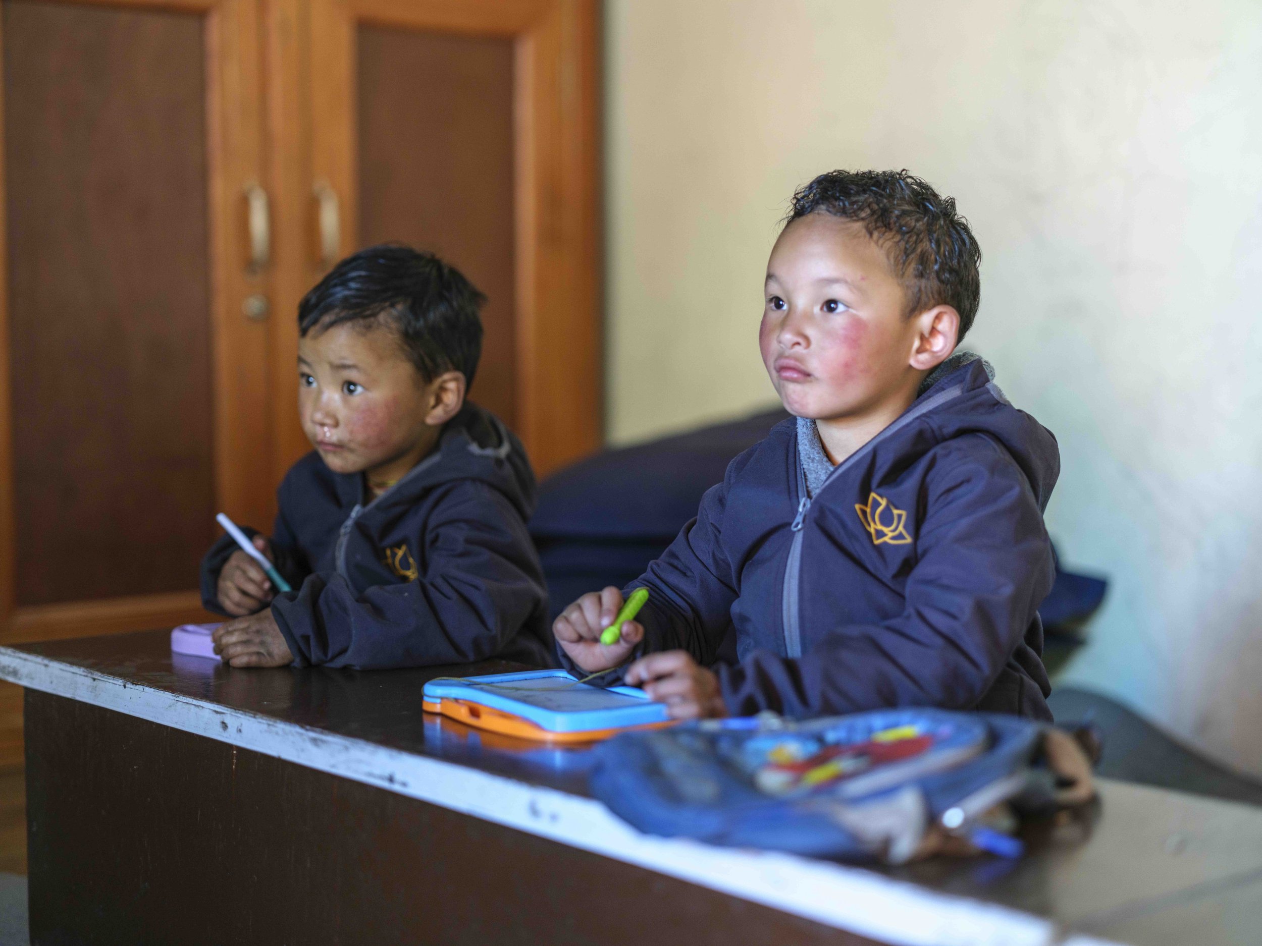 Two young boys sitting at a desk with colorful notebooks and pens, focusing attentively, in a classroom setting.