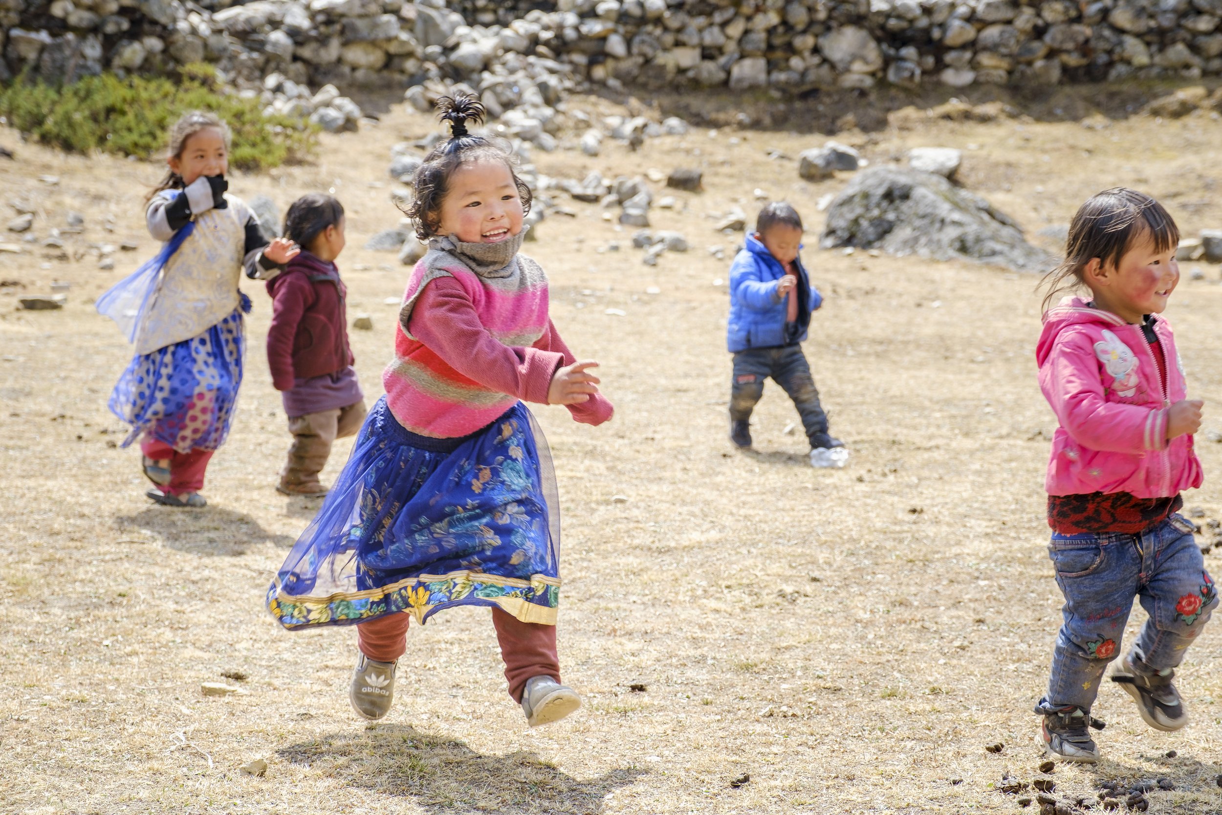 Group of young children running and playing outdoors on a dirt surface with rocks and sparse vegetation in the background.