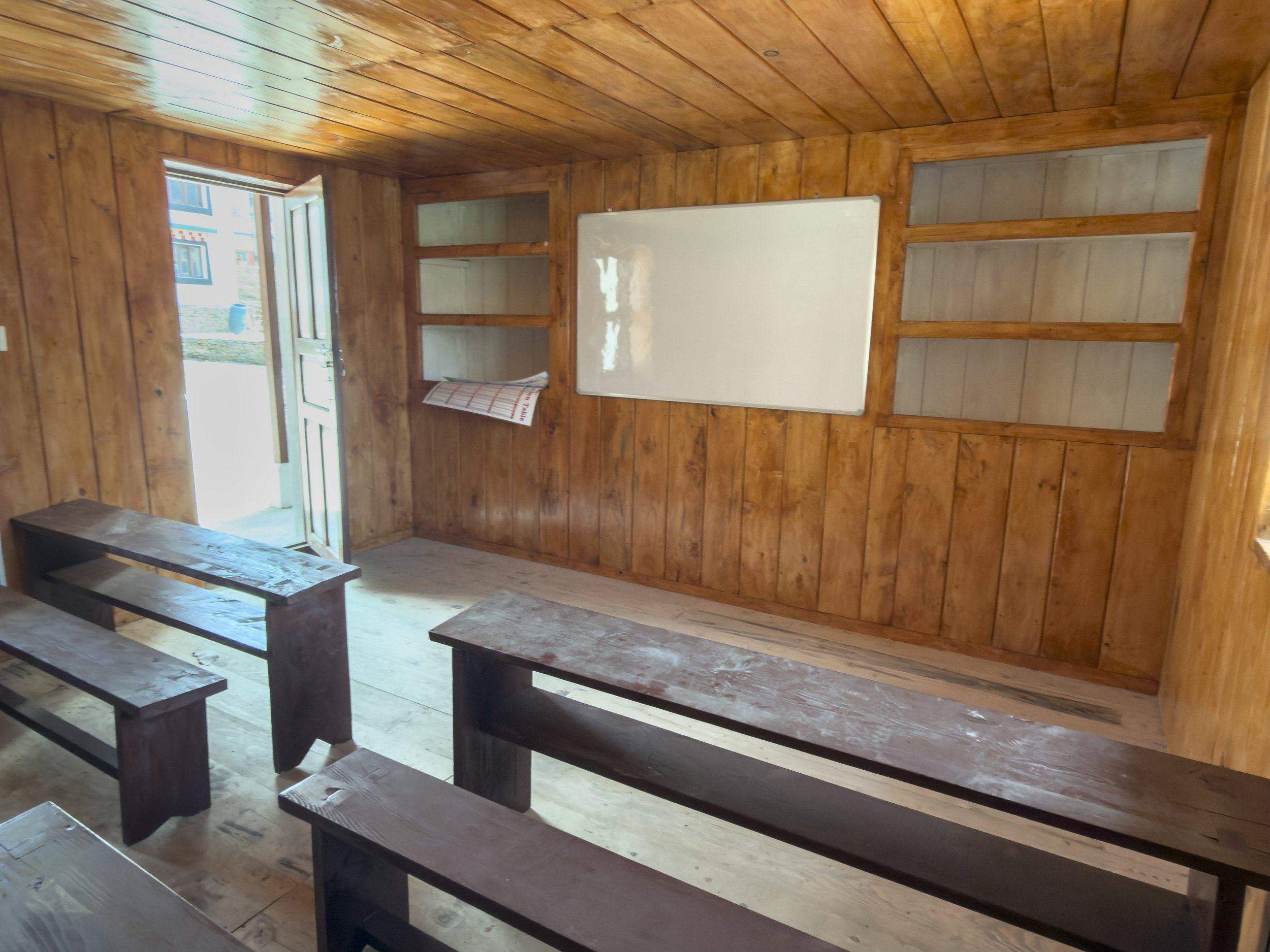 Interior of a wooden cabin with wood-paneled walls and ceiling, a whiteboard mounted on the wall, shelves, and a window with an open door showing an outdoor scene.