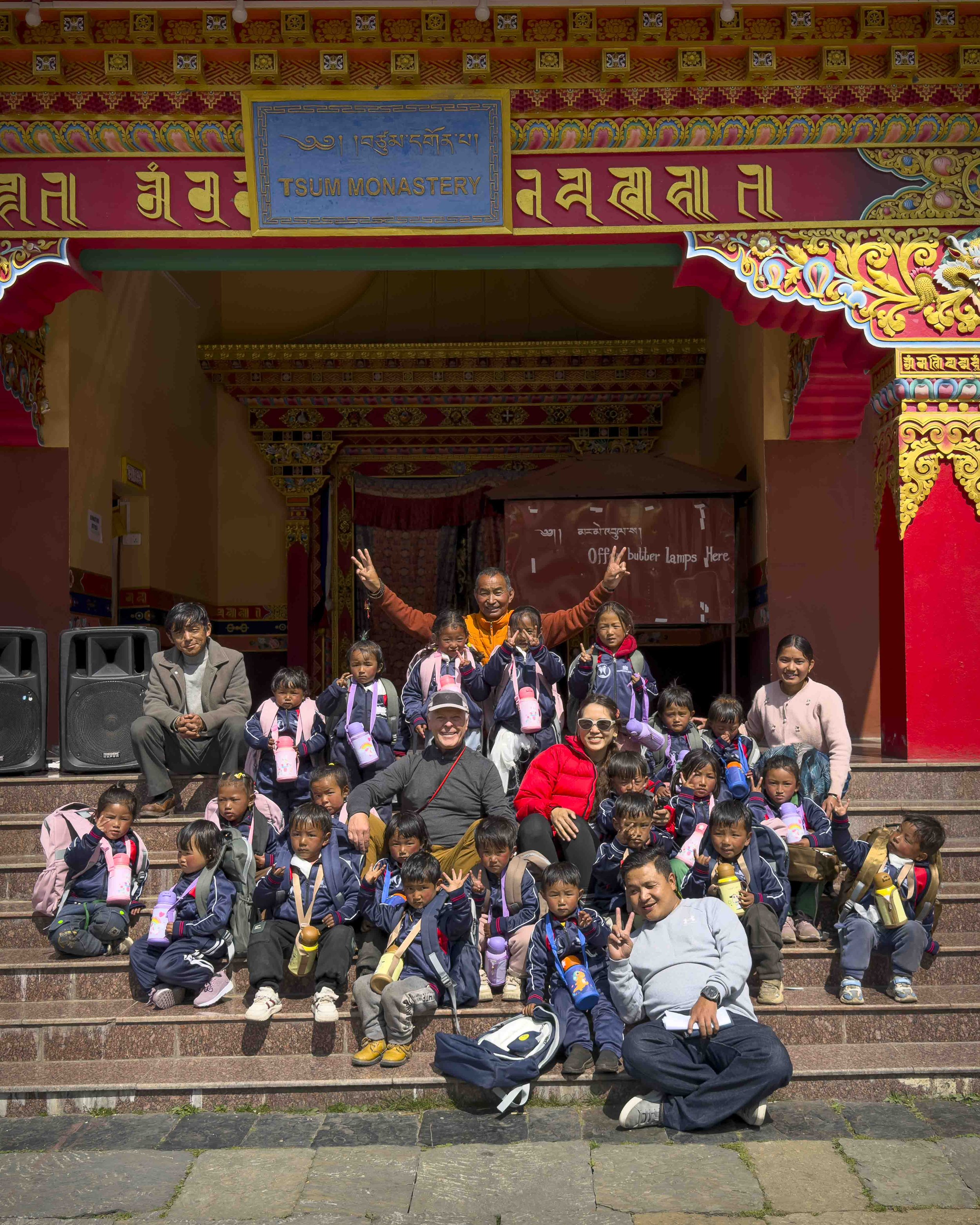 A group of children, adults, and seniors sitting on steps in front of the entrance to Tsum Monastery, a colorful traditional Buddhist building with intricate decorative patterns and a sign with the monastery's name.